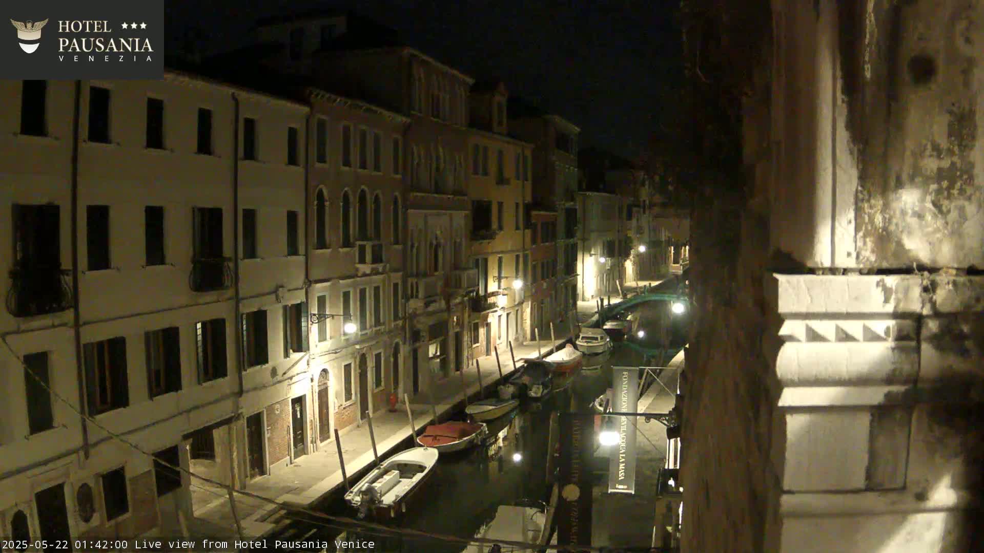 A nighttime view of a narrow canal in Venice, lined with buildings, with several boats moored along its edge.