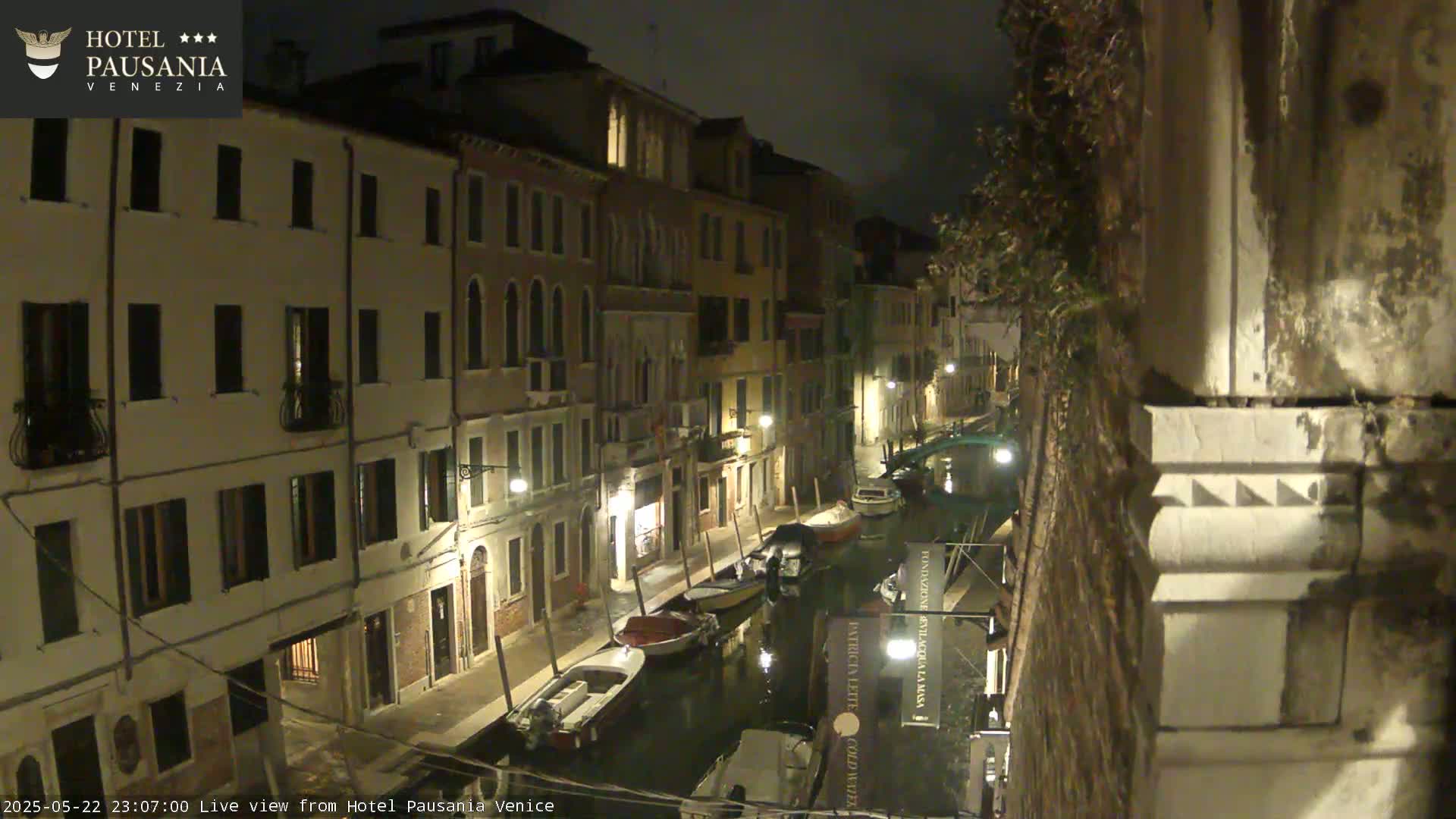 A nighttime view of a narrow canal in Venice, lined with buildings, with several boats moored along its edge.