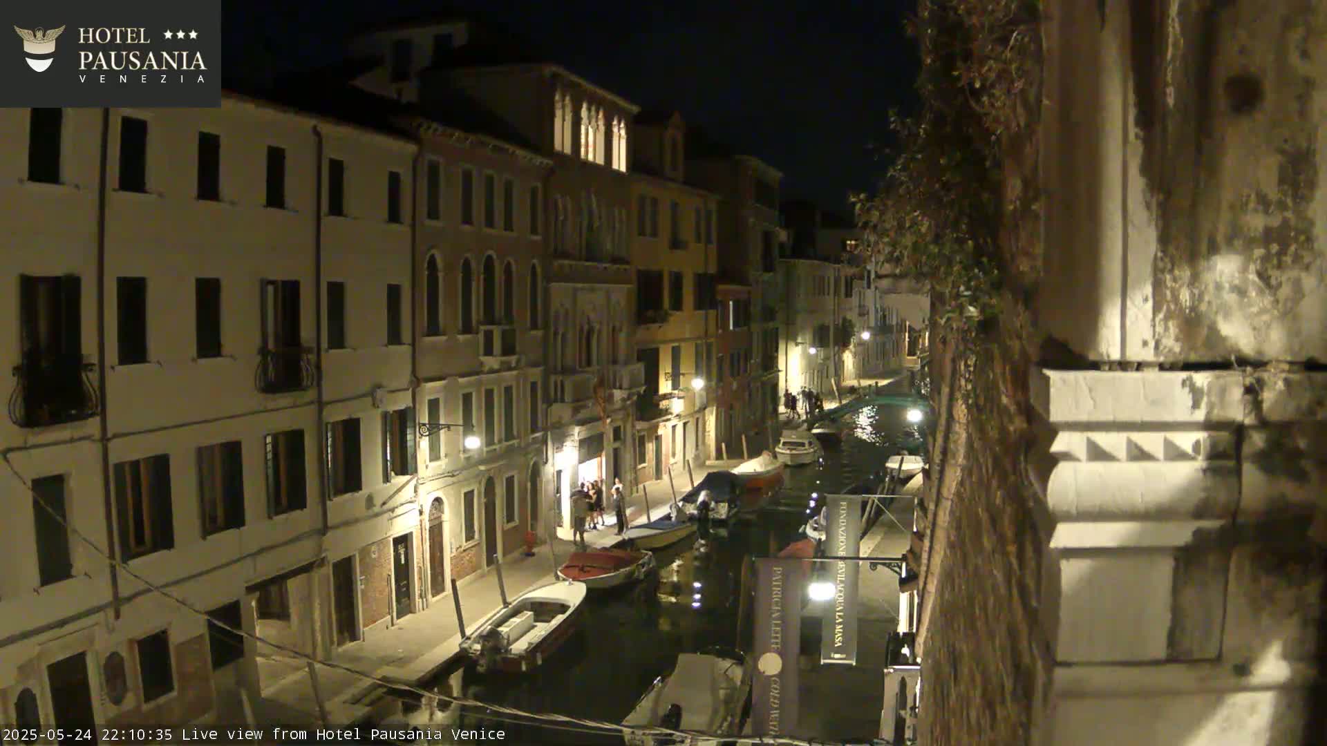 A nighttime view of a narrow canal in Venice, with several boats moored along its banks and buildings lining both sides of the waterway, illuminated by streetlights.