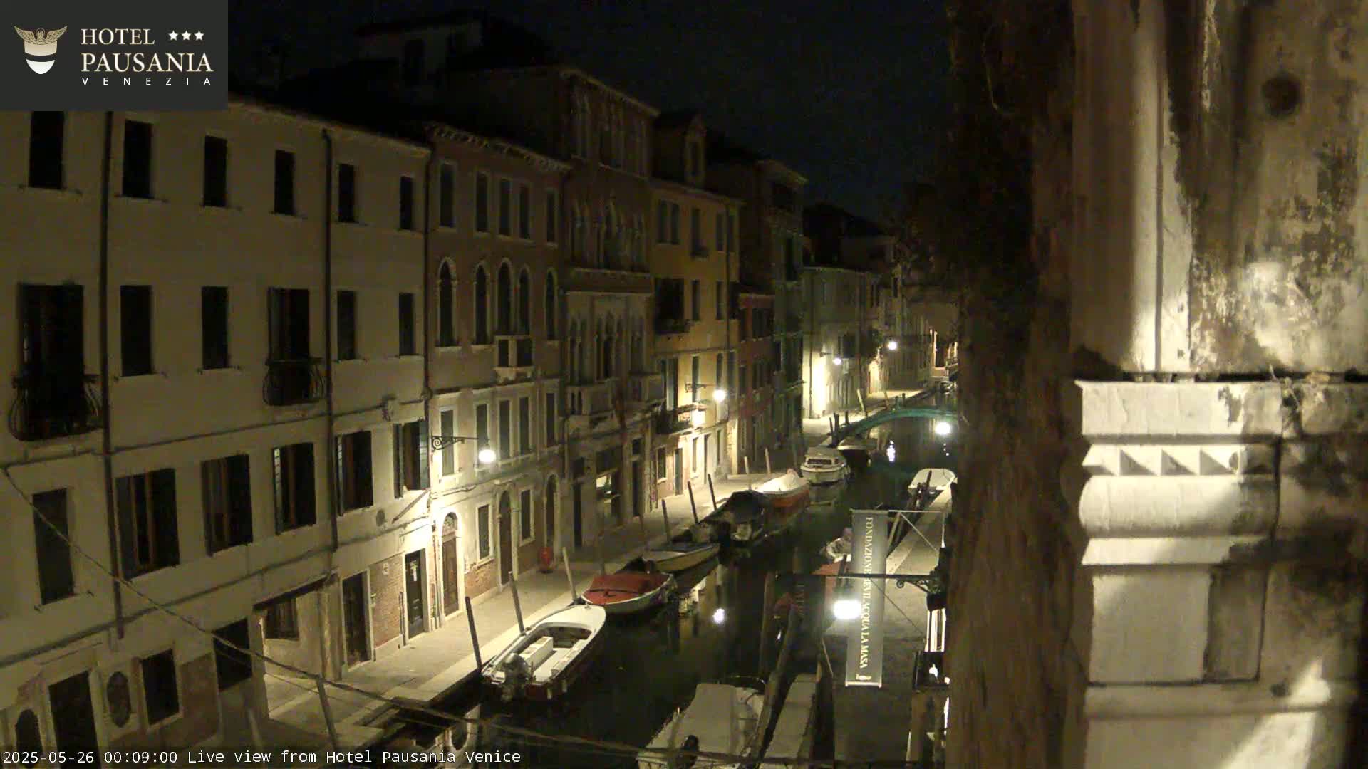 A nighttime view of a narrow canal in Venice, Italy, lined with illuminated buildings and several moored boats.