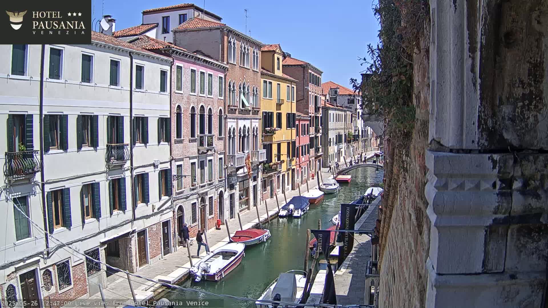 A sunny day in Venice shows a narrow canal lined with colorful buildings and several motorboats moored along its banks.