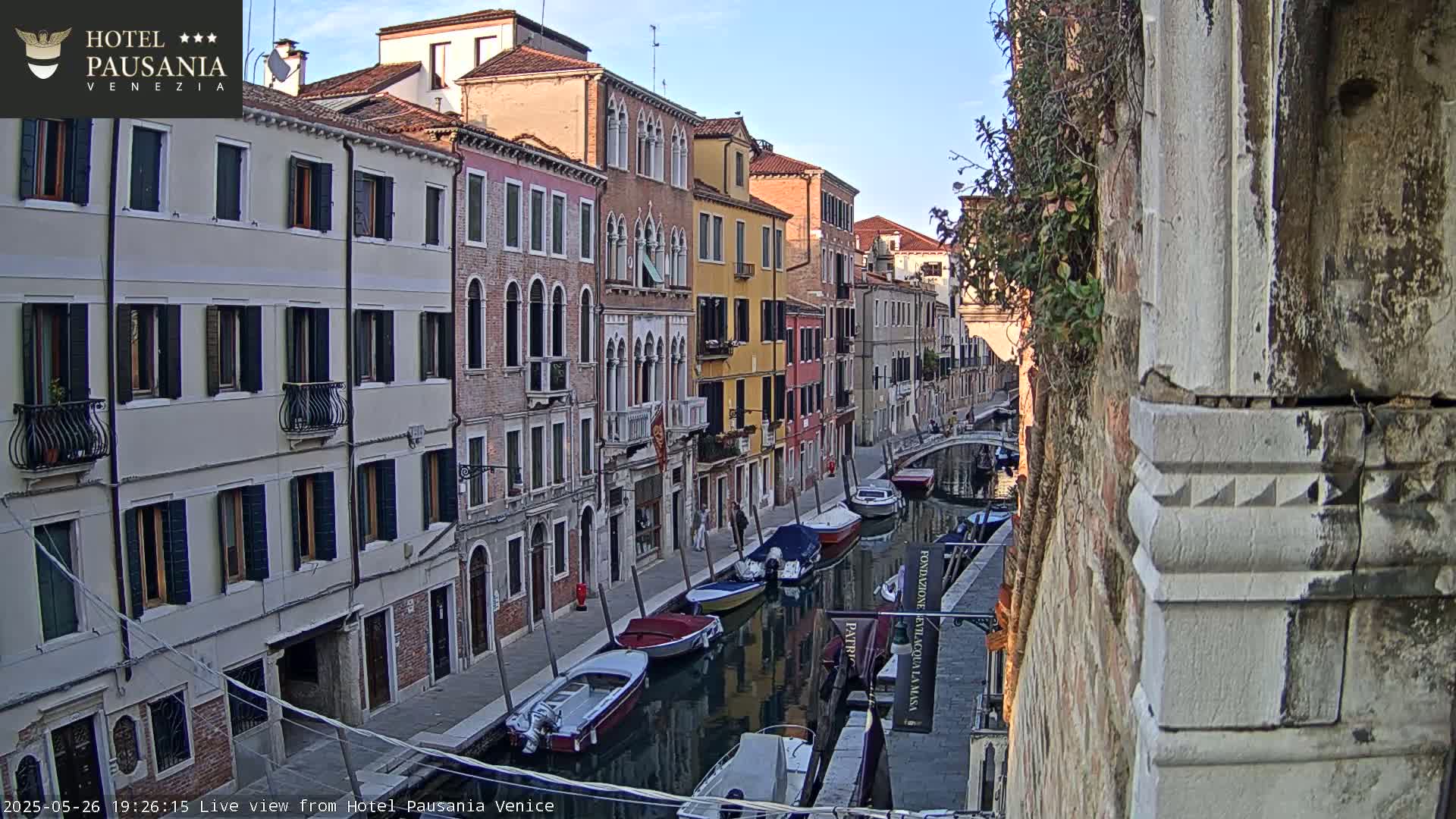 A narrow Venetian canal is lined with colorful buildings under a clear, bright sky, several boats are moored along the waterway.
