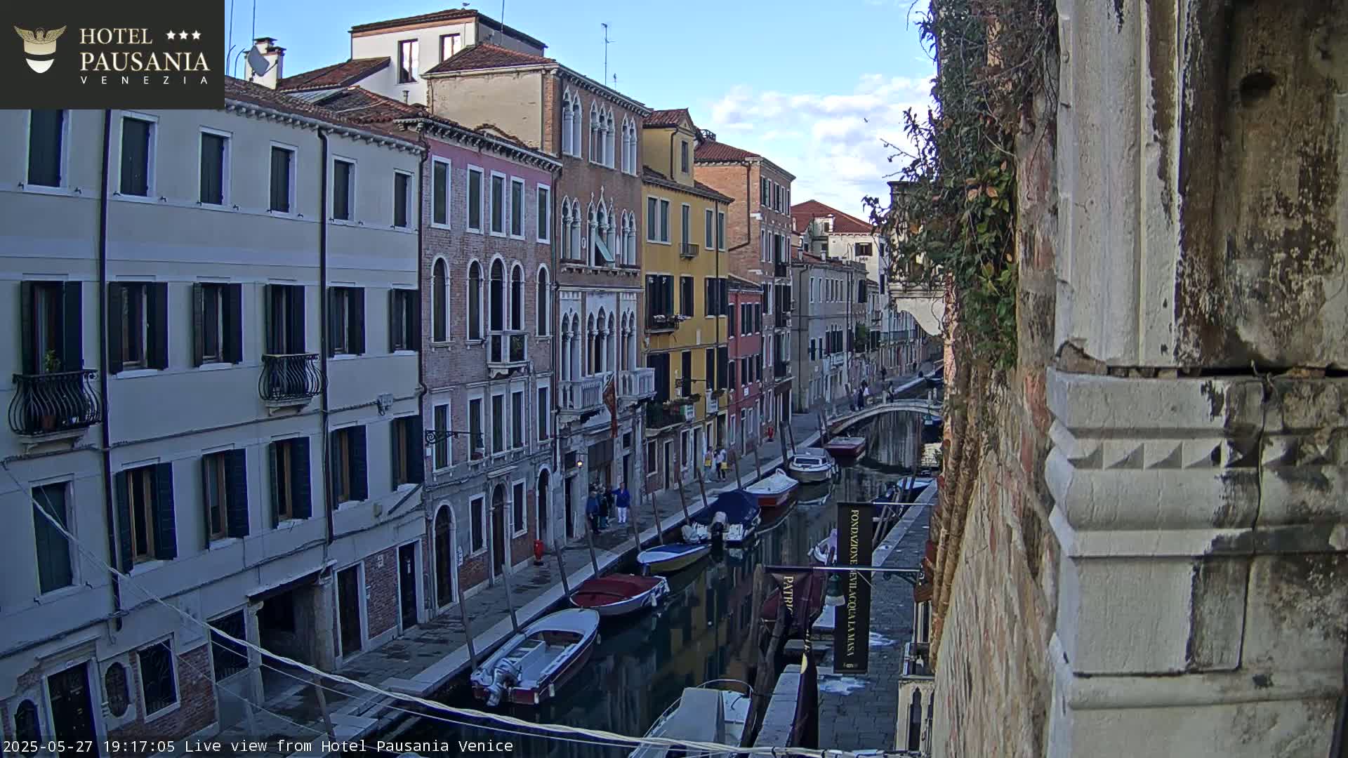 A narrow Venetian canal, under a clear sky, is lined with colorful buildings and several moored boats.