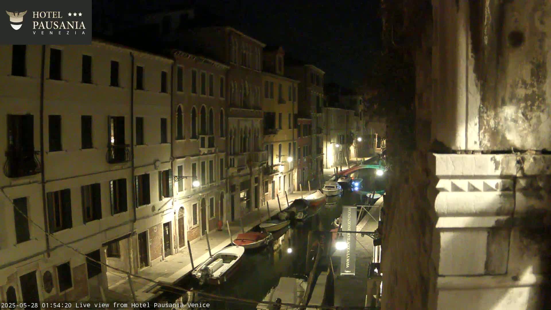 A nighttime view of a narrow canal in Venice, lined with illuminated buildings and several small boats moored along its banks.
