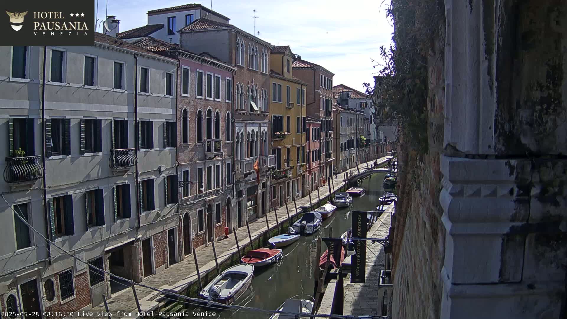 A sunny day in Venice shows a narrow canal lined with colorful buildings and several moored boats.
