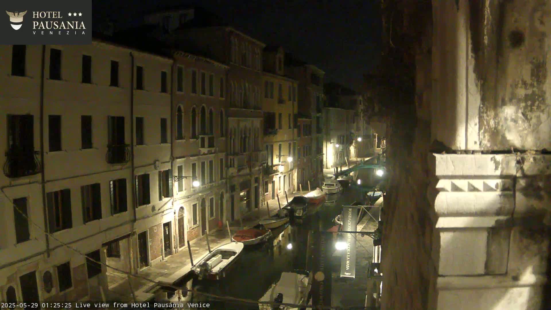 A nighttime view of a narrow canal in Venice, lined with illuminated buildings and several moored boats.