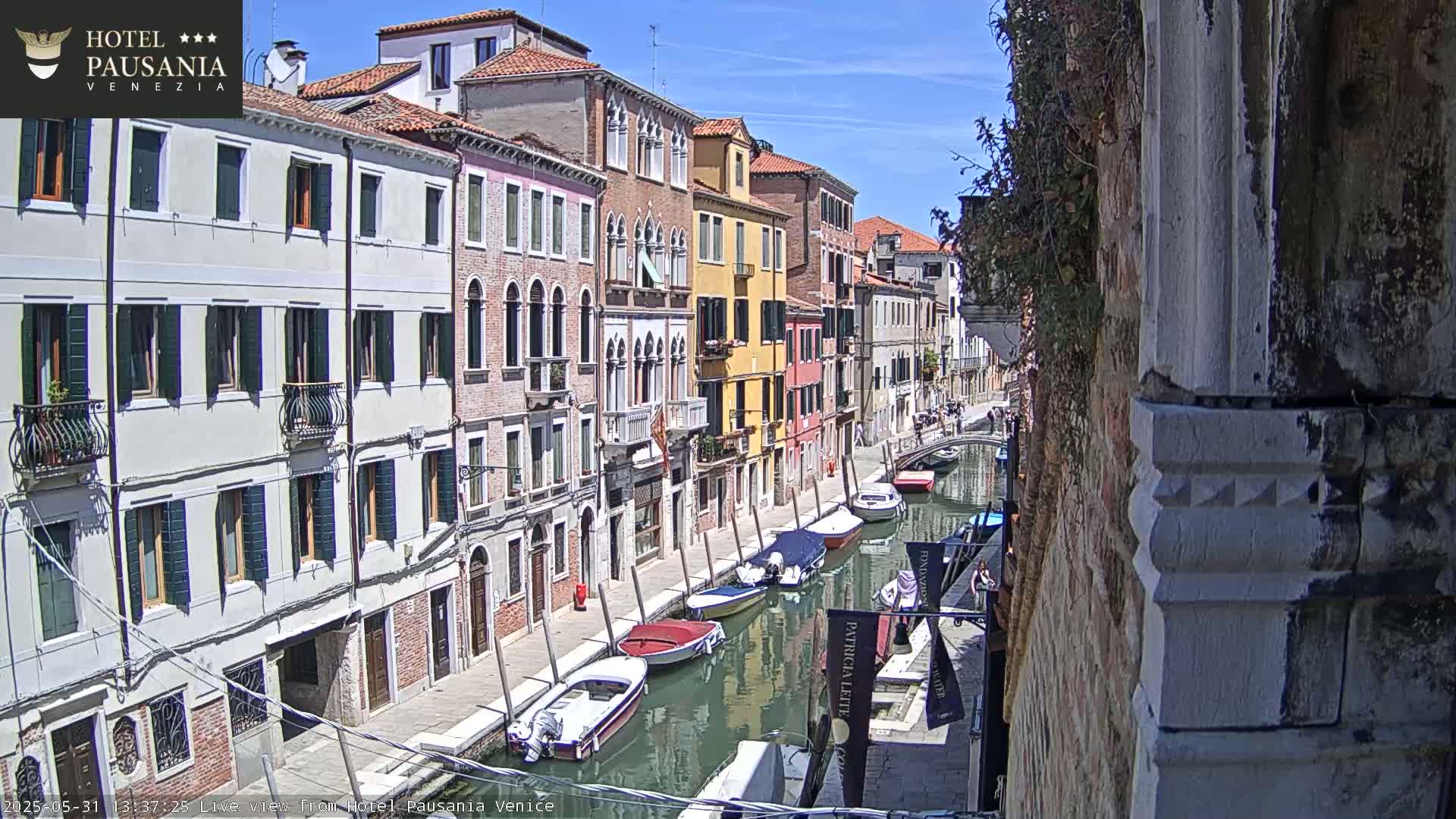 A sunny day in Venice shows a narrow canal lined with colorful buildings and several moored boats.
