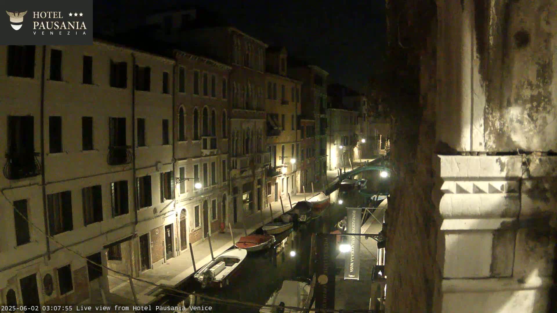 A nighttime view of a narrow canal in Venice, lined with buildings and several moored boats, under a clear, dark sky.