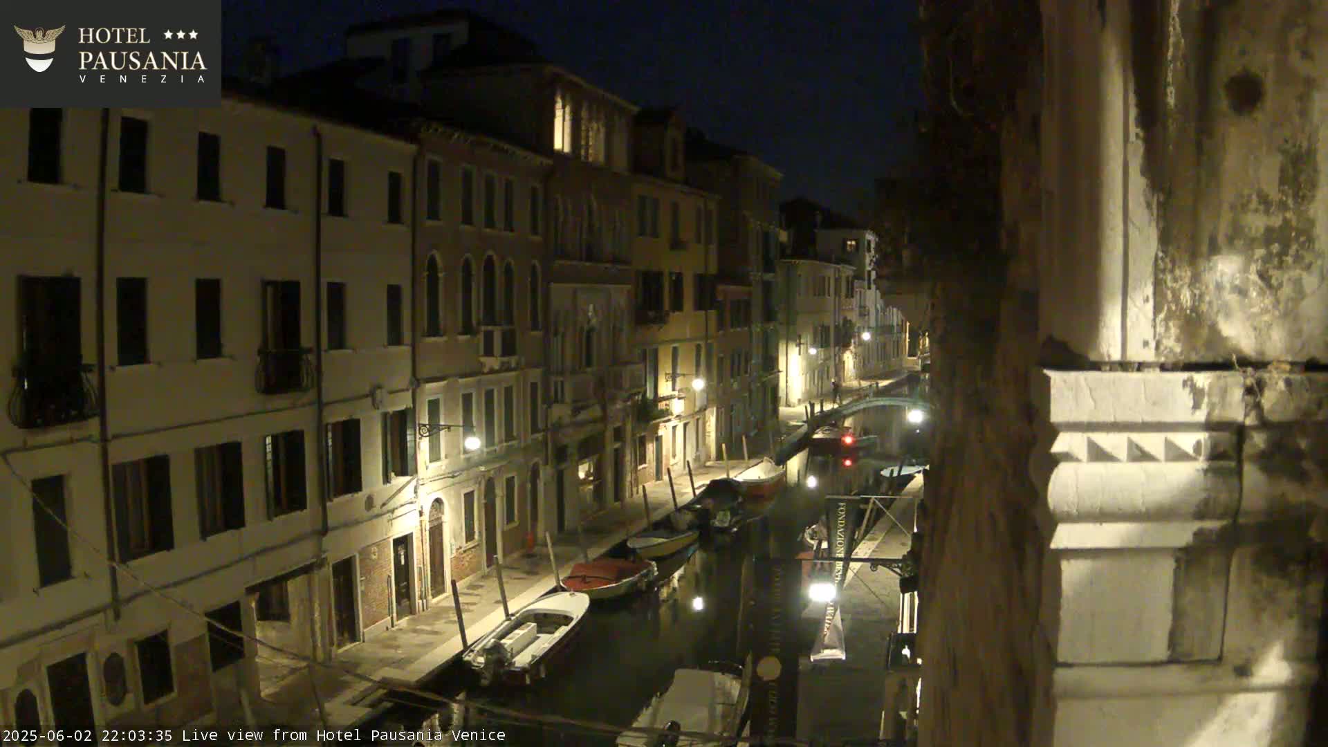 A nighttime view of a narrow canal in Venice, Italy, lined with illuminated buildings and a few moored boats.