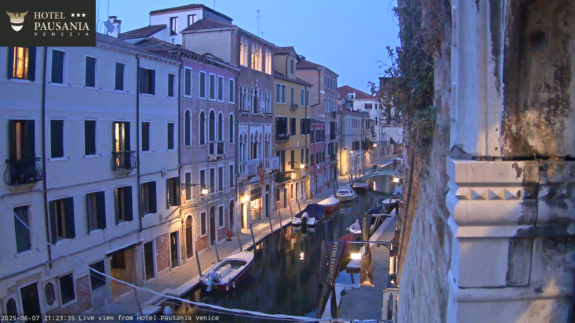 A twilight view of a narrow canal in Venice, lined with tall, illuminated buildings and several moored boats.
