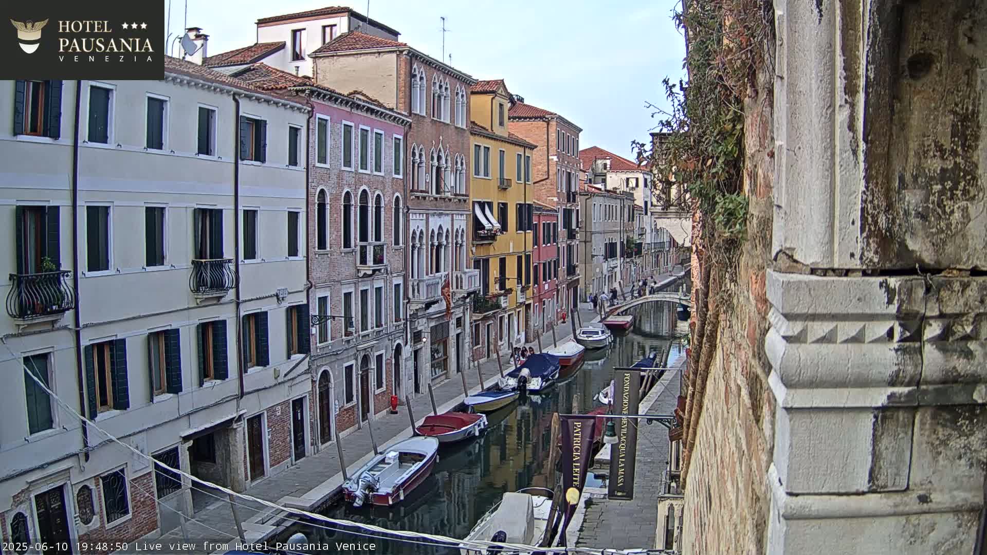 A narrow canal in Venice, under a mostly clear sky, is lined with colorful buildings and several boats are moored along its banks.