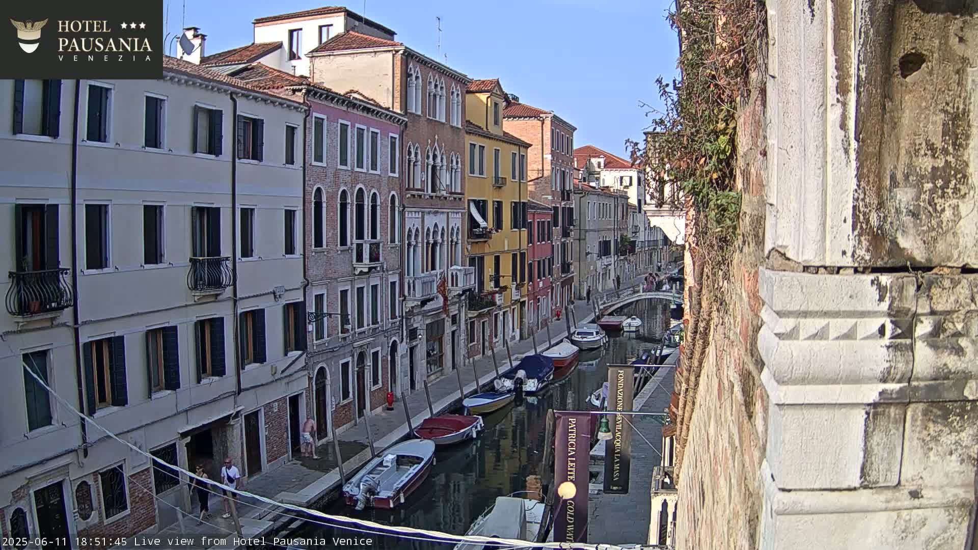 A narrow Venetian canal is lined with colorful buildings under a sunny sky, and several boats are moored along its banks.