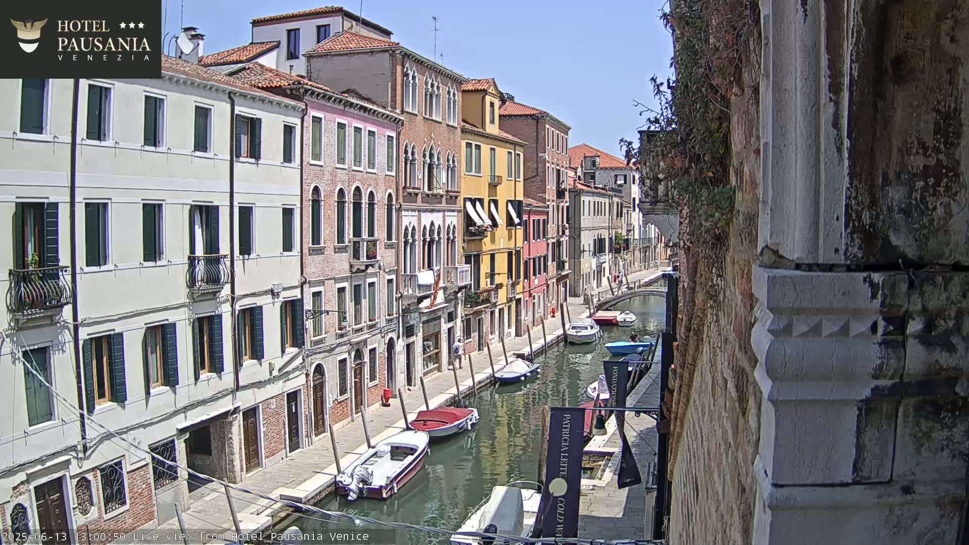 A sunny day in Venice shows a narrow canal lined with colorful buildings and several small boats moored along the waterway.