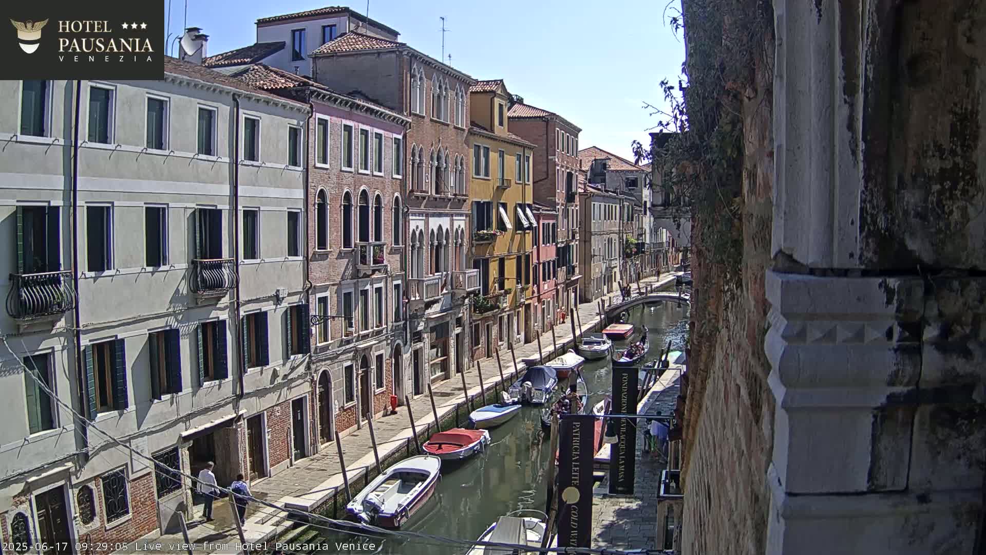 A sunny day in Venice shows a narrow canal lined with colorful buildings and several boats moored along the waterway.