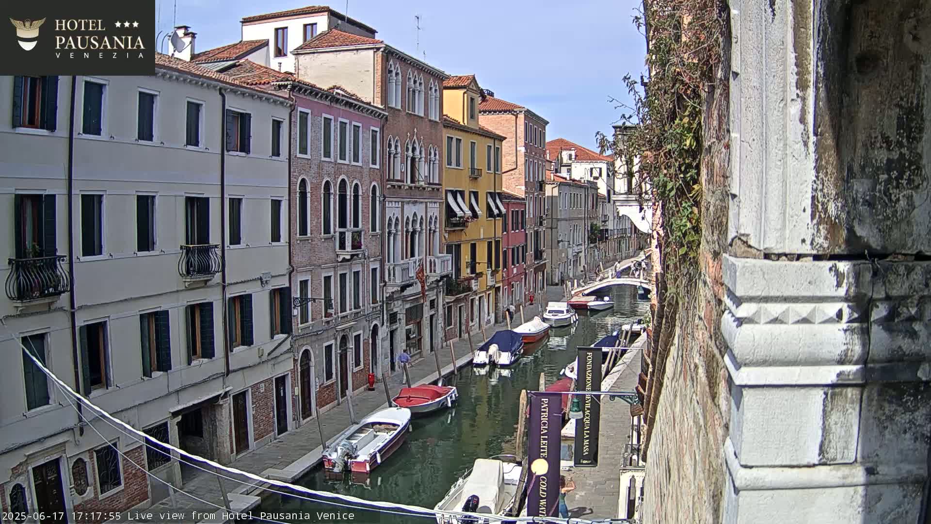 A sunny day in Venice, showing a narrow canal lined with colorful buildings and several boats moored along its sides.