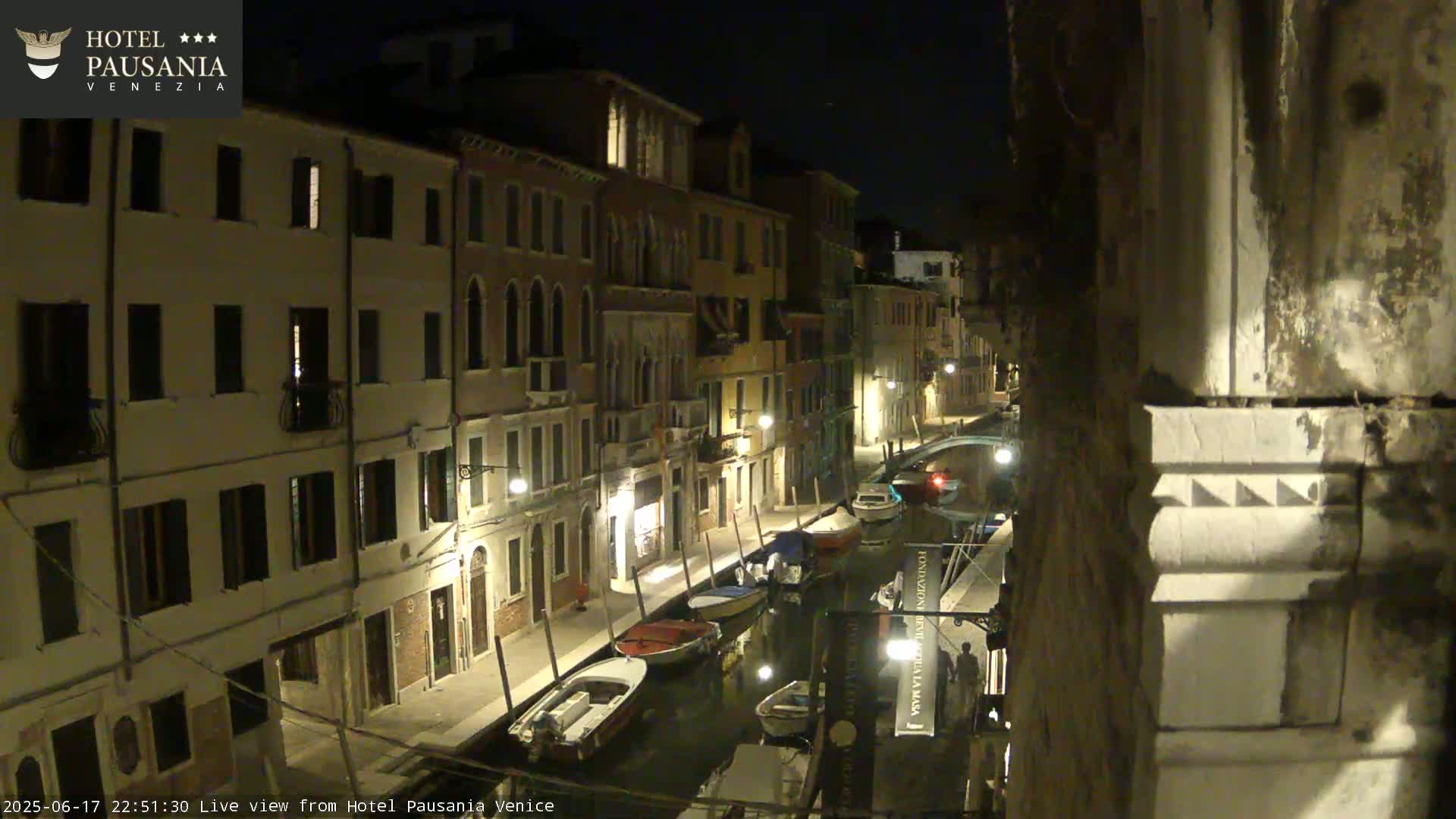 A nighttime view of a narrow canal in Venice, with several boats moored along its banks and illuminated buildings lining the waterway.