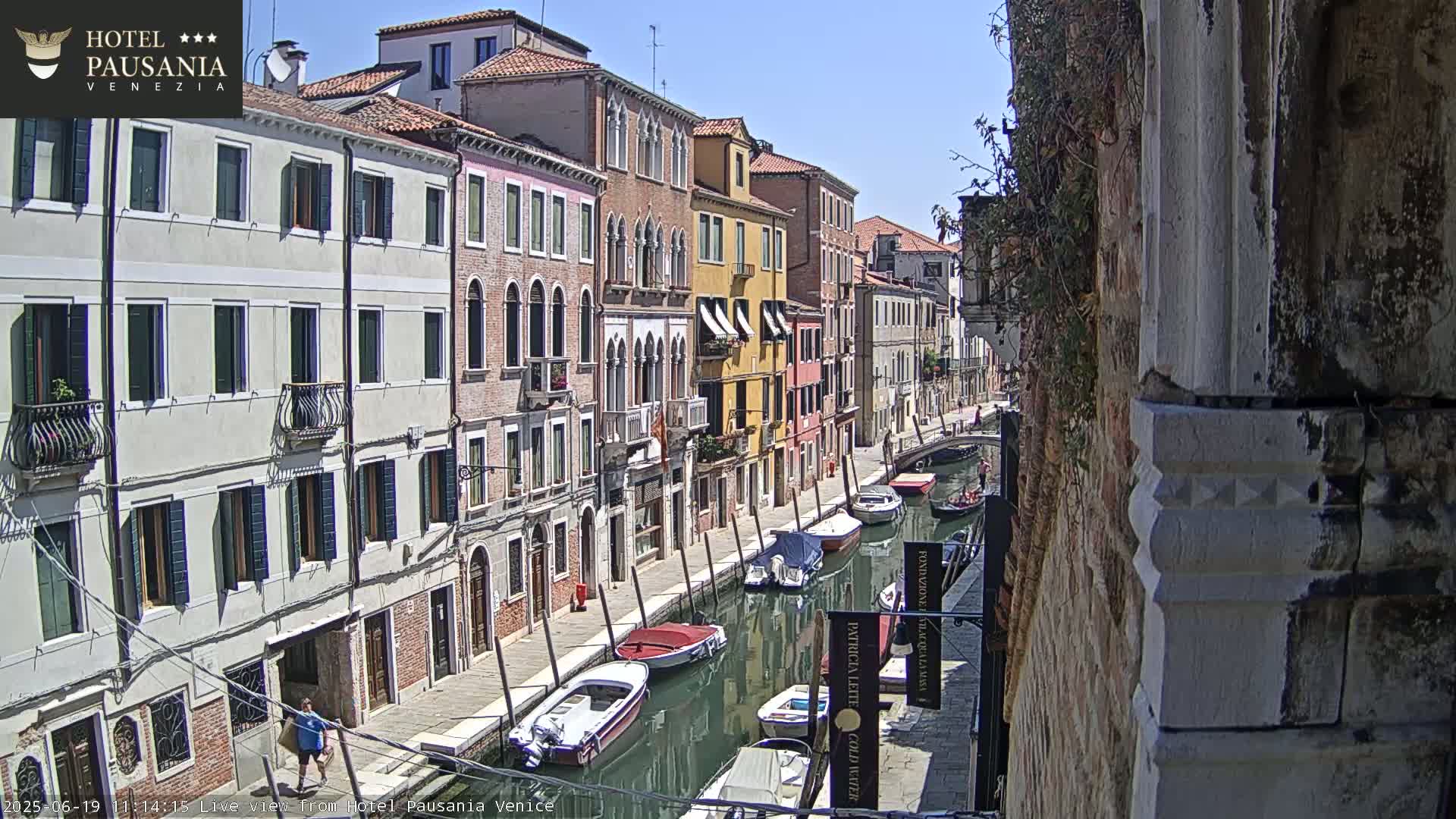 A sunny day in Venice shows a narrow canal lined with colorful buildings, several boats moored along its sides, and a person walking along the canal path.