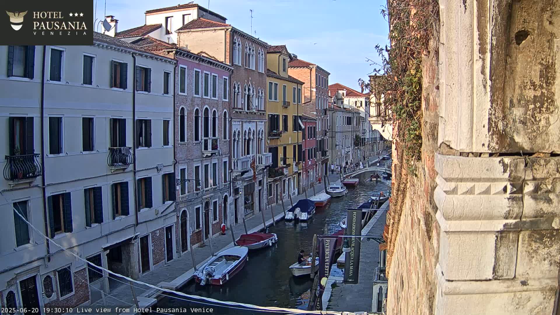 A narrow canal in Venice, Italy, lined with tall, historic buildings under a clear, sunny sky, is filled with several moored motorboats.
