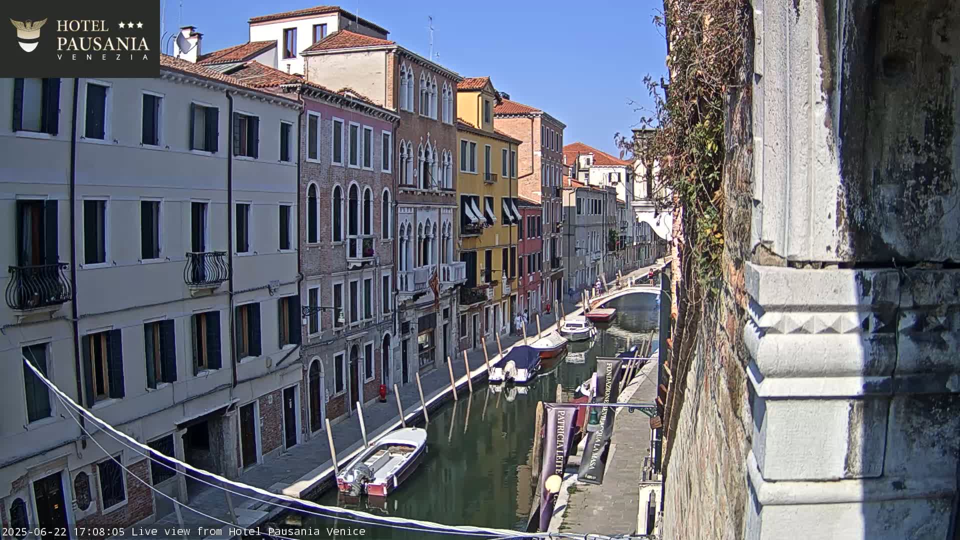 A sunny Venetian canal scene with colorful buildings lining its banks, several small boats moored along the waterway.