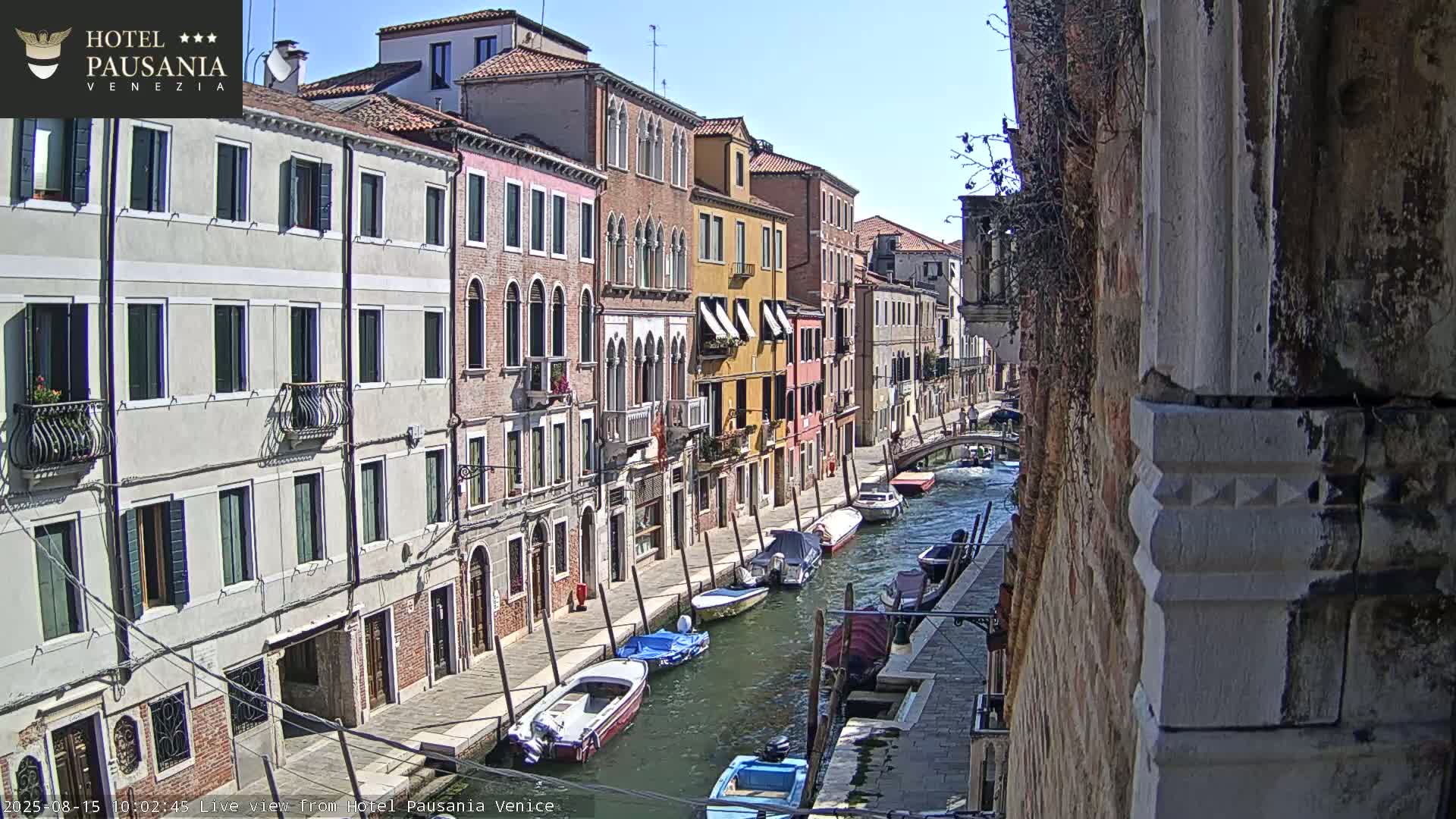 A sunny day in Venice shows a narrow canal lined with colorful buildings and several moored boats.