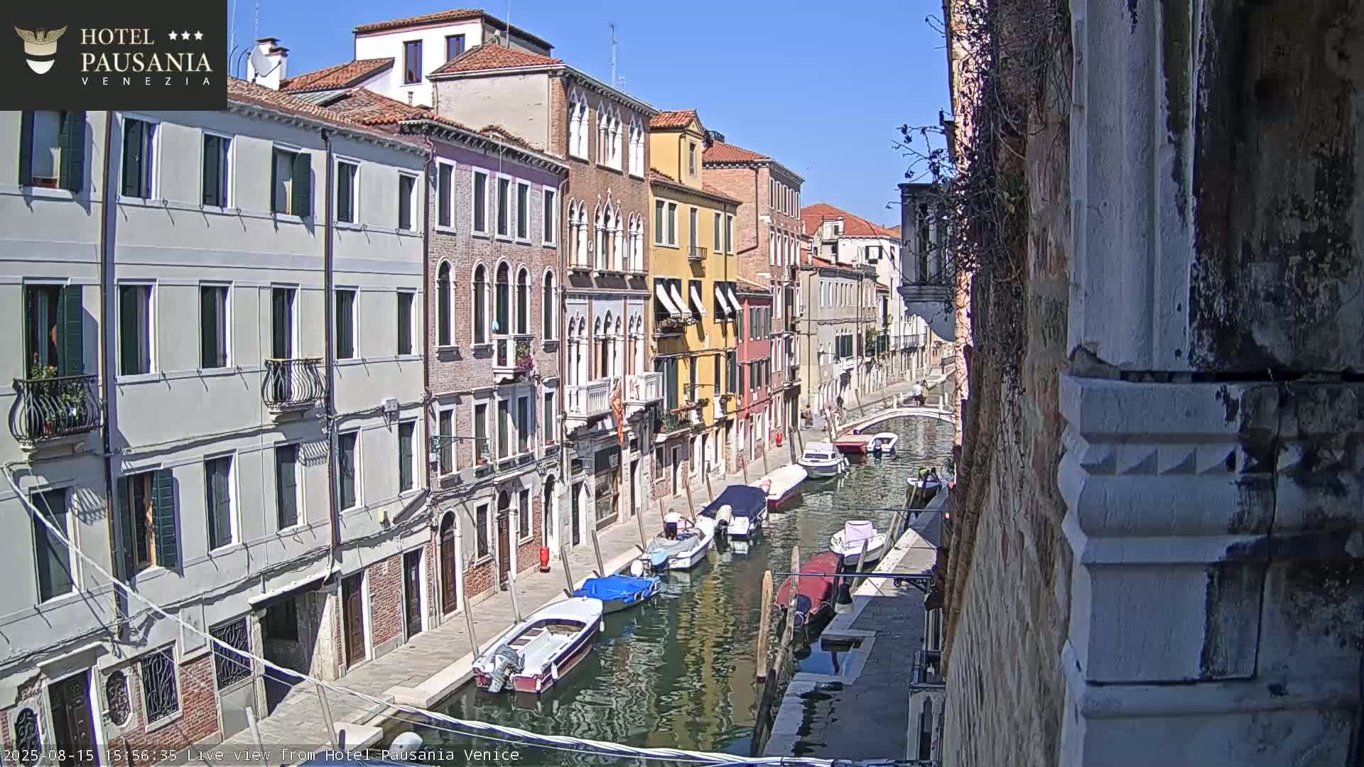 A sunny day in Venice shows a narrow canal lined with colorful buildings and several moored boats.