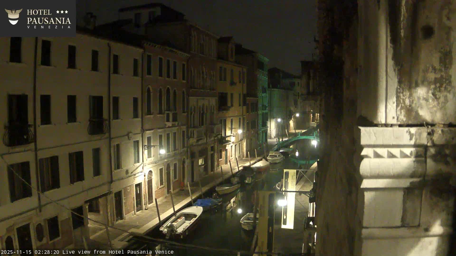 The image shows a clear, dark night scene of a narrow canal in Venice, lined with illuminated historic buildings and moored boats, with a small arched bridge visible in the distance.