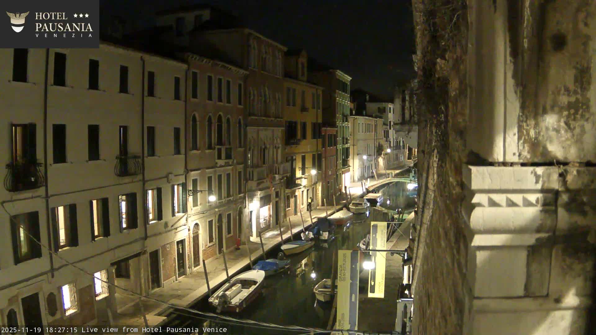 The image shows a clear, dark night scene of a narrow canal in Venice, lined with illuminated historic buildings and moored boats, with a small arched bridge visible in the distance.