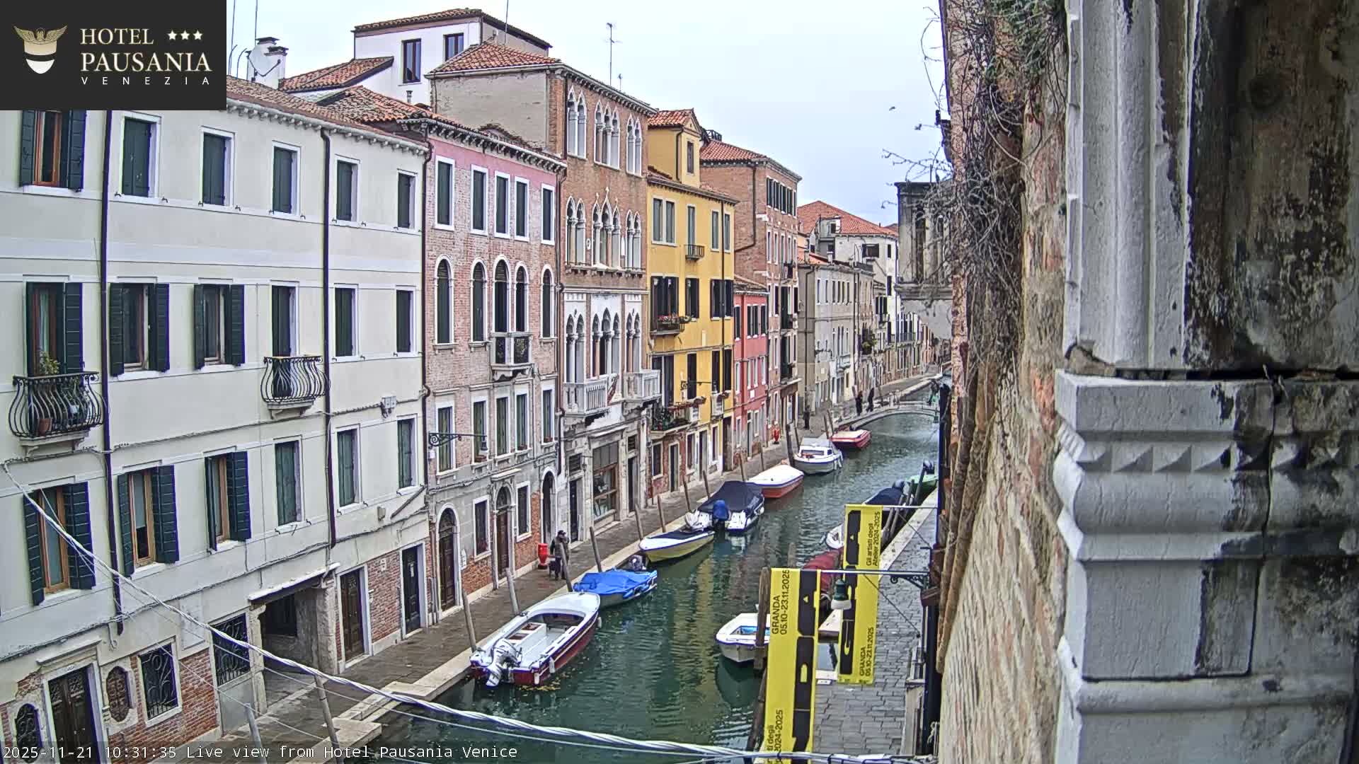 A tranquil Venetian canal is lined with a colorful array of historic buildings, featuring numerous moored boats and a distant arched bridge, all under an overcast sky.