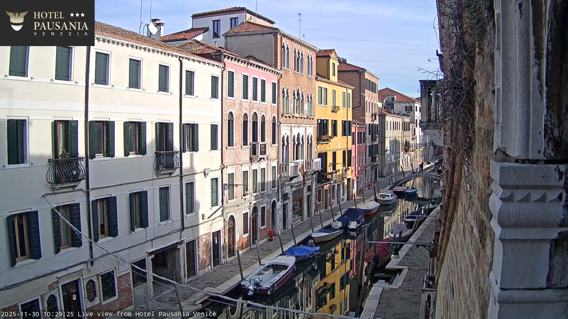 A sunny day in Venice reveals a narrow canal lined with historic, multi-colored buildings, their reflections shimmering in the water alongside numerous moored boats, all beneath a clear blue sky.