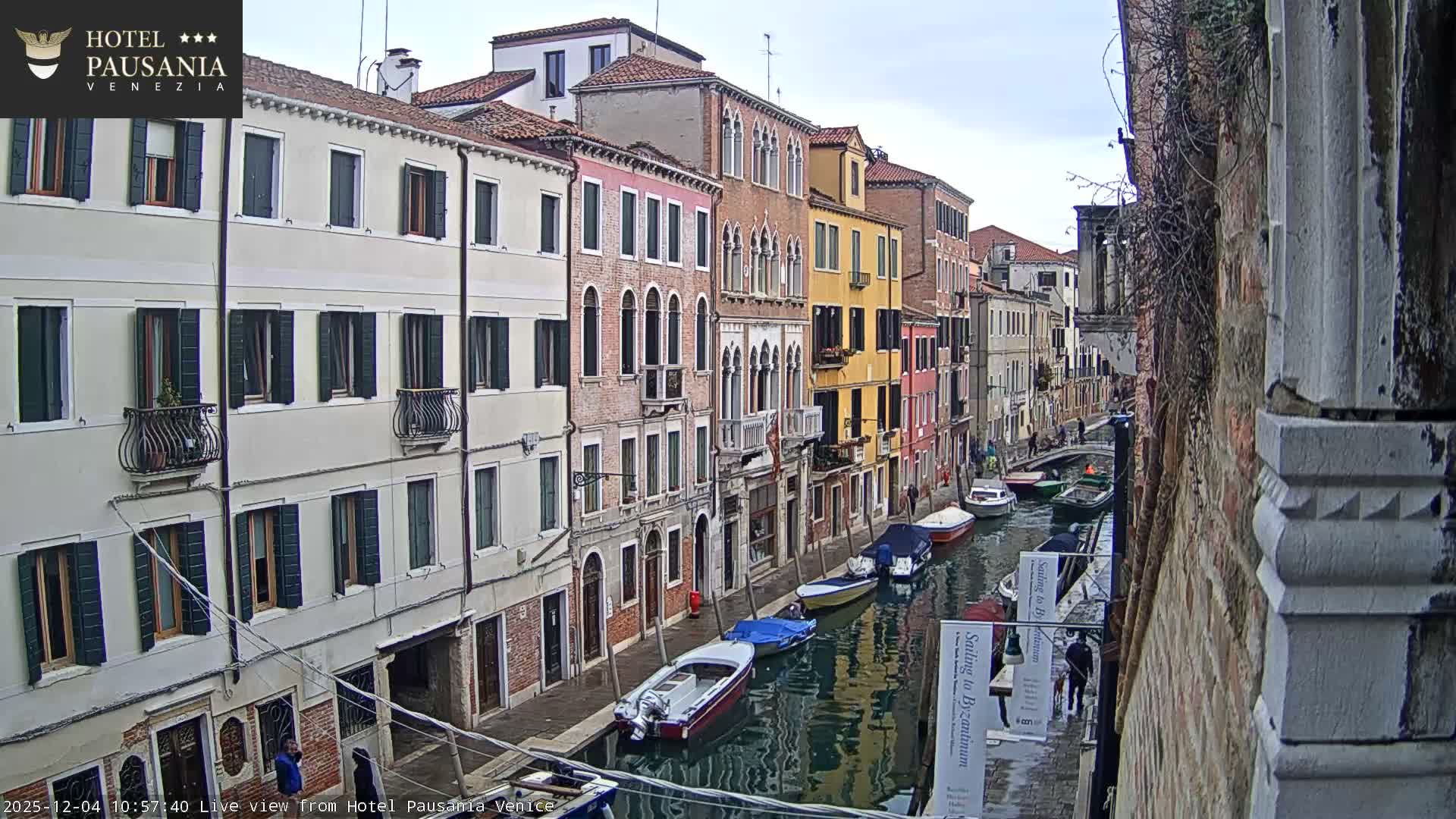 An elevated view captures a winding Venetian canal filled with several boats, flanked by a dense array of colorful historic buildings and bridged walkways with people, all under a dull, cloudy sky.