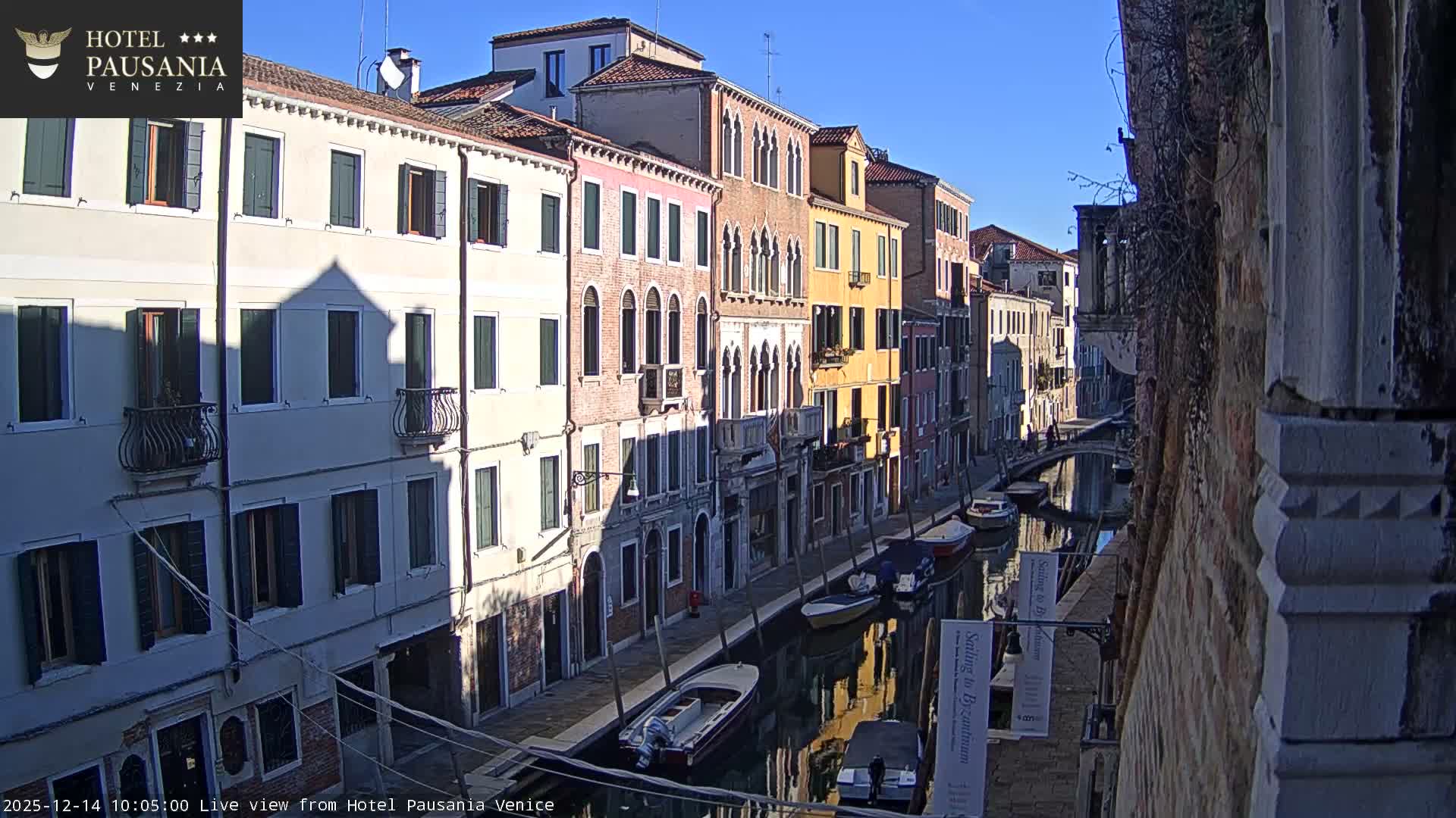 An elevated view captures a winding Venetian canal filled with several boats, flanked by a dense array of colorful historic buildings and bridged walkways with people, all under a dull, cloudy sky.