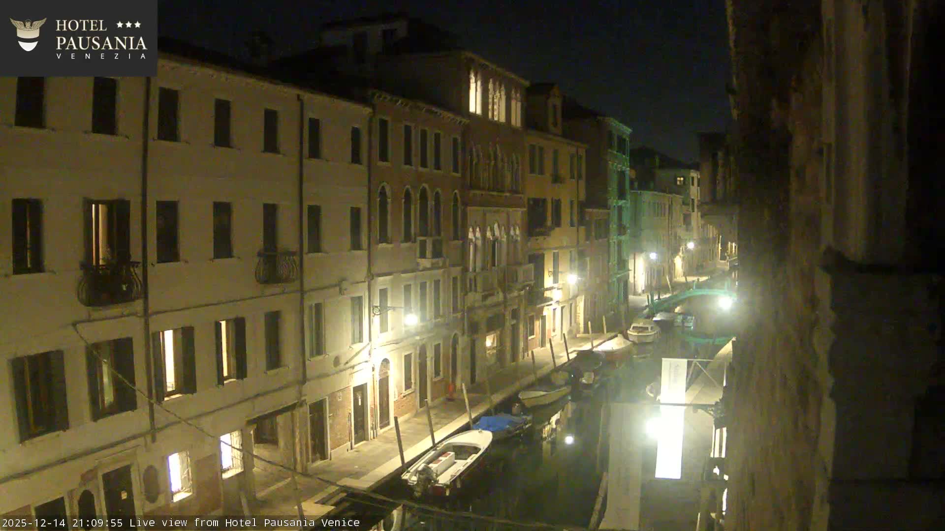 An elevated view captures a winding Venetian canal filled with several boats, flanked by a dense array of colorful historic buildings and bridged walkways with people, all under a dull, cloudy sky.