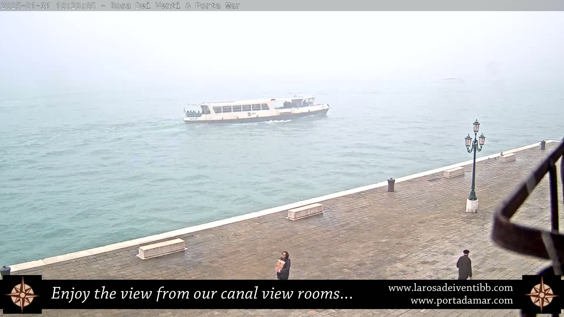 A ferry boat travels on a waterway under foggy conditions, alongside a paved waterfront area with two people standing.