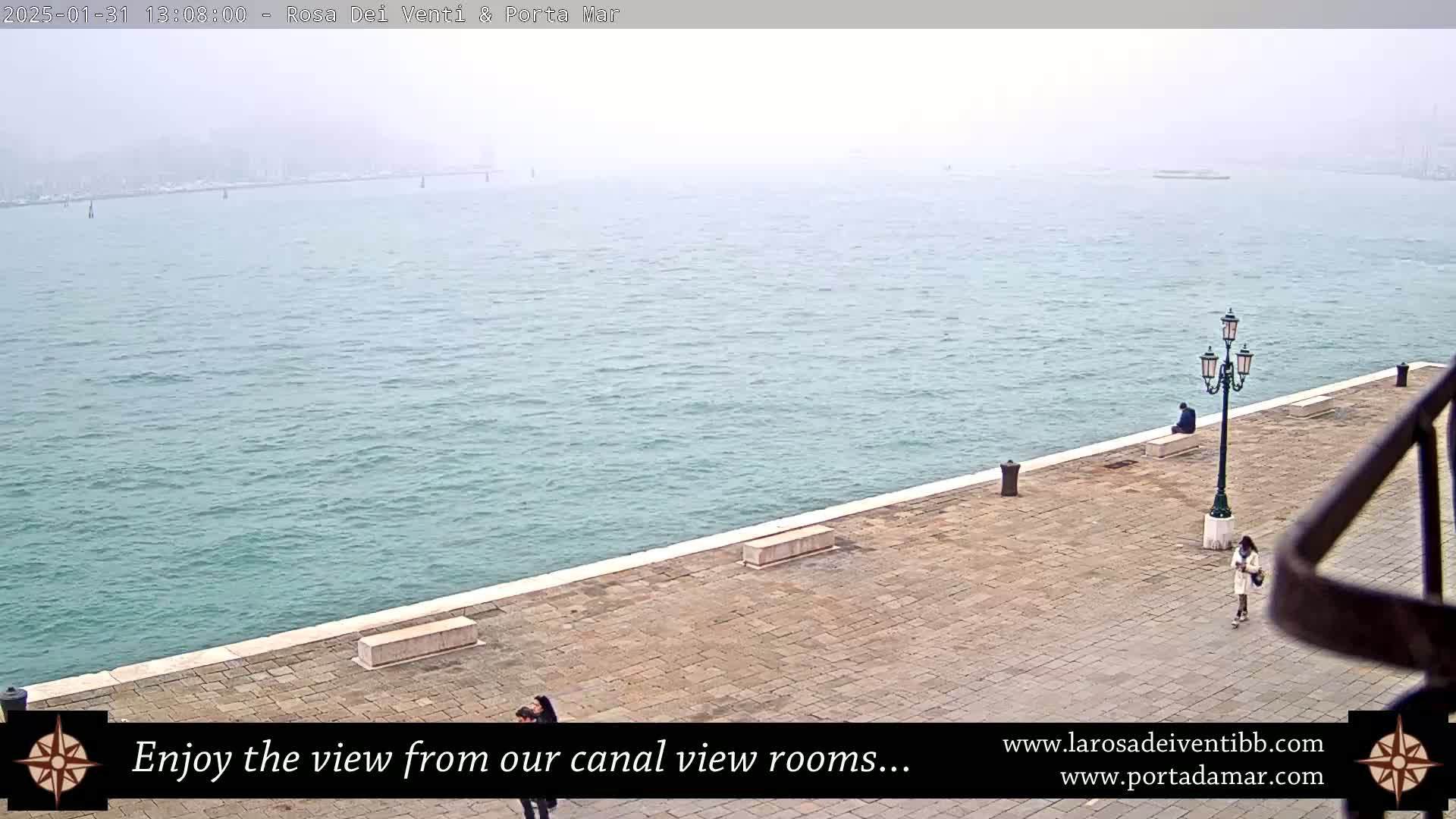 A hazy, overcast day reveals a calm body of water alongside a stone walkway with a few people walking and sitting.