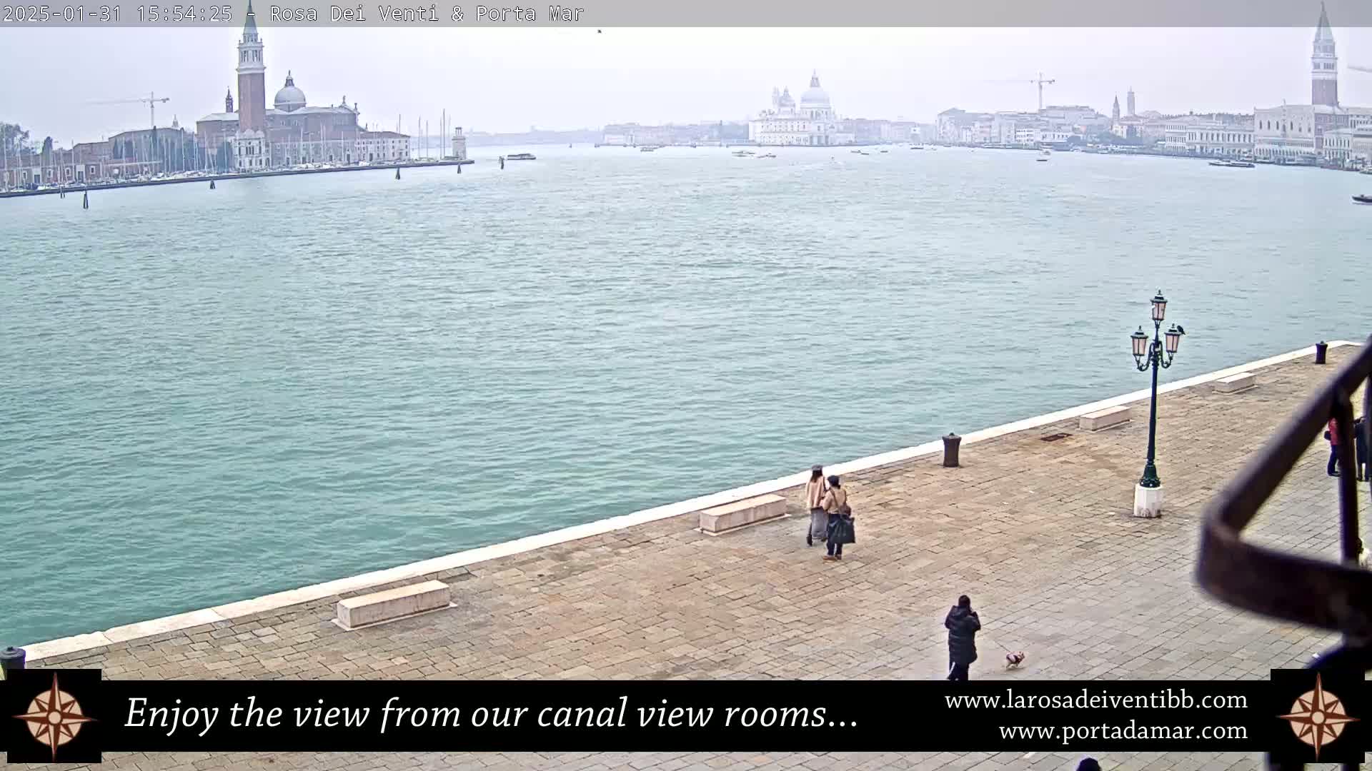 A wide shot of a calm canal in Venice, Italy on an overcast day, shows a paved walkway with several people and a lamppost, with buildings and churches visible in the distance.