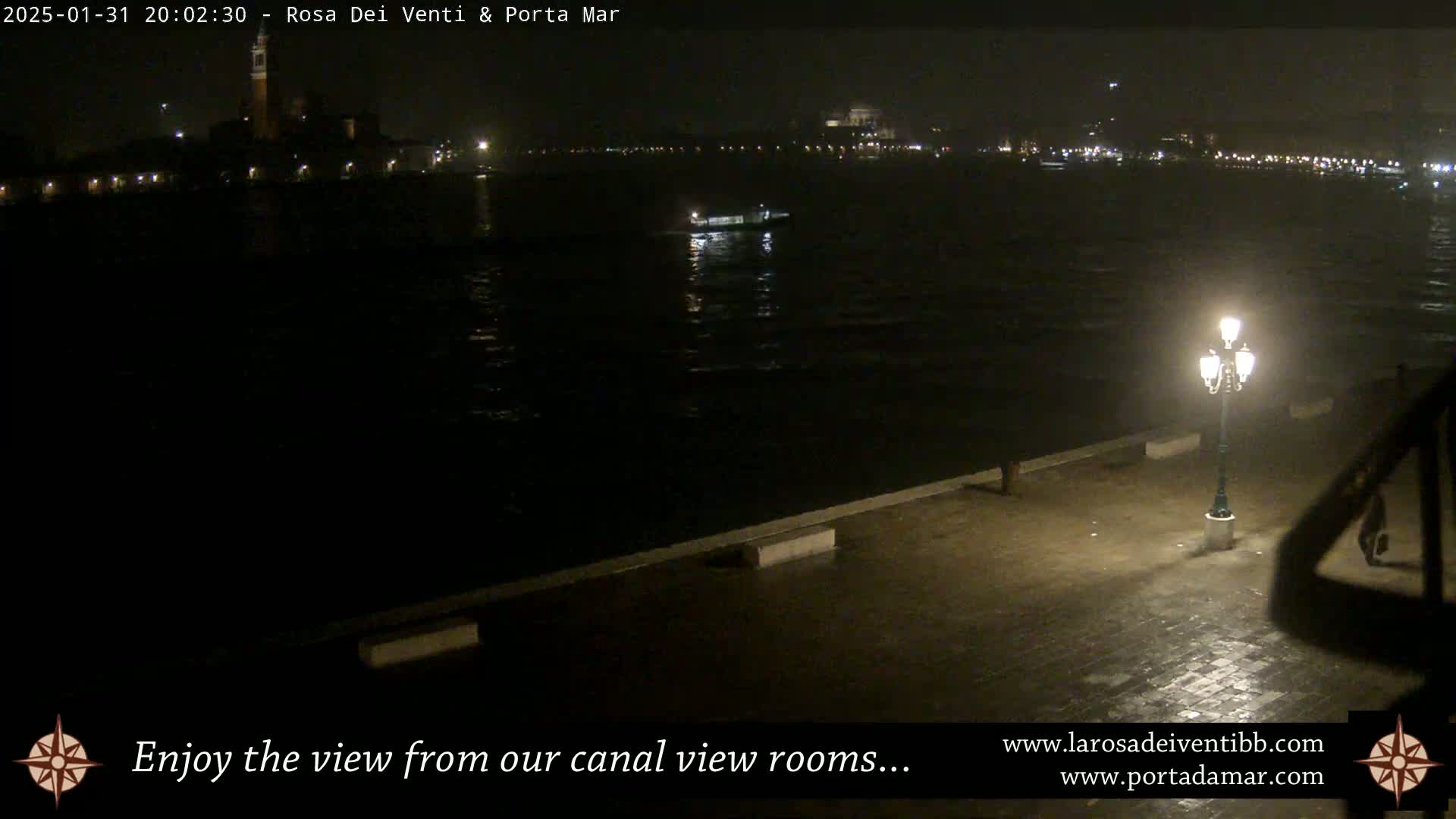A nighttime view of a dark canal, a small boat on the water, and a single lit lamppost on a paved waterfront.