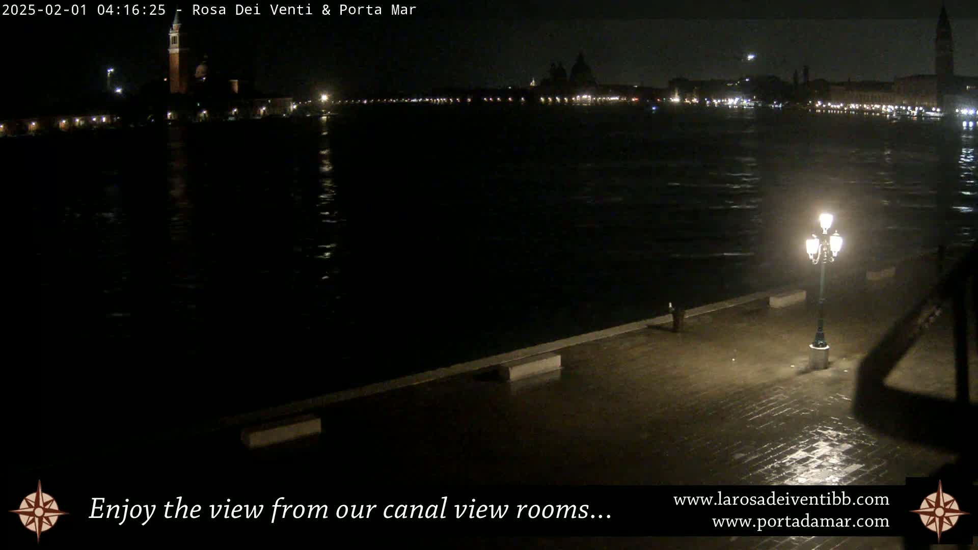 A nighttime view across a dark canal shows the lights of a city across the water, with a single lit lamppost on a wet, paved surface in the foreground.