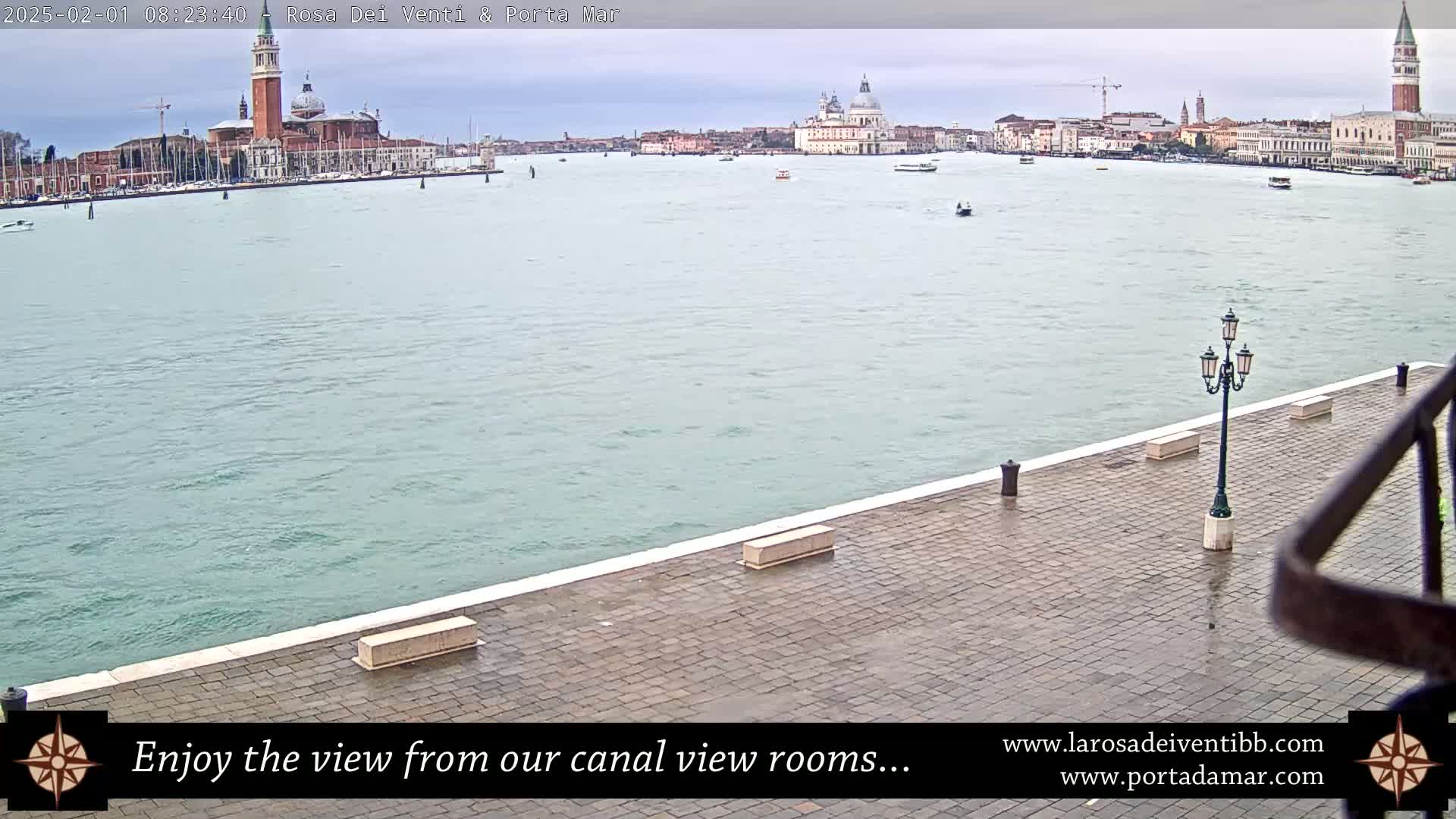 A wet stone plaza overlooks a calm canal, with a distant view of buildings and a bell tower under an overcast sky.