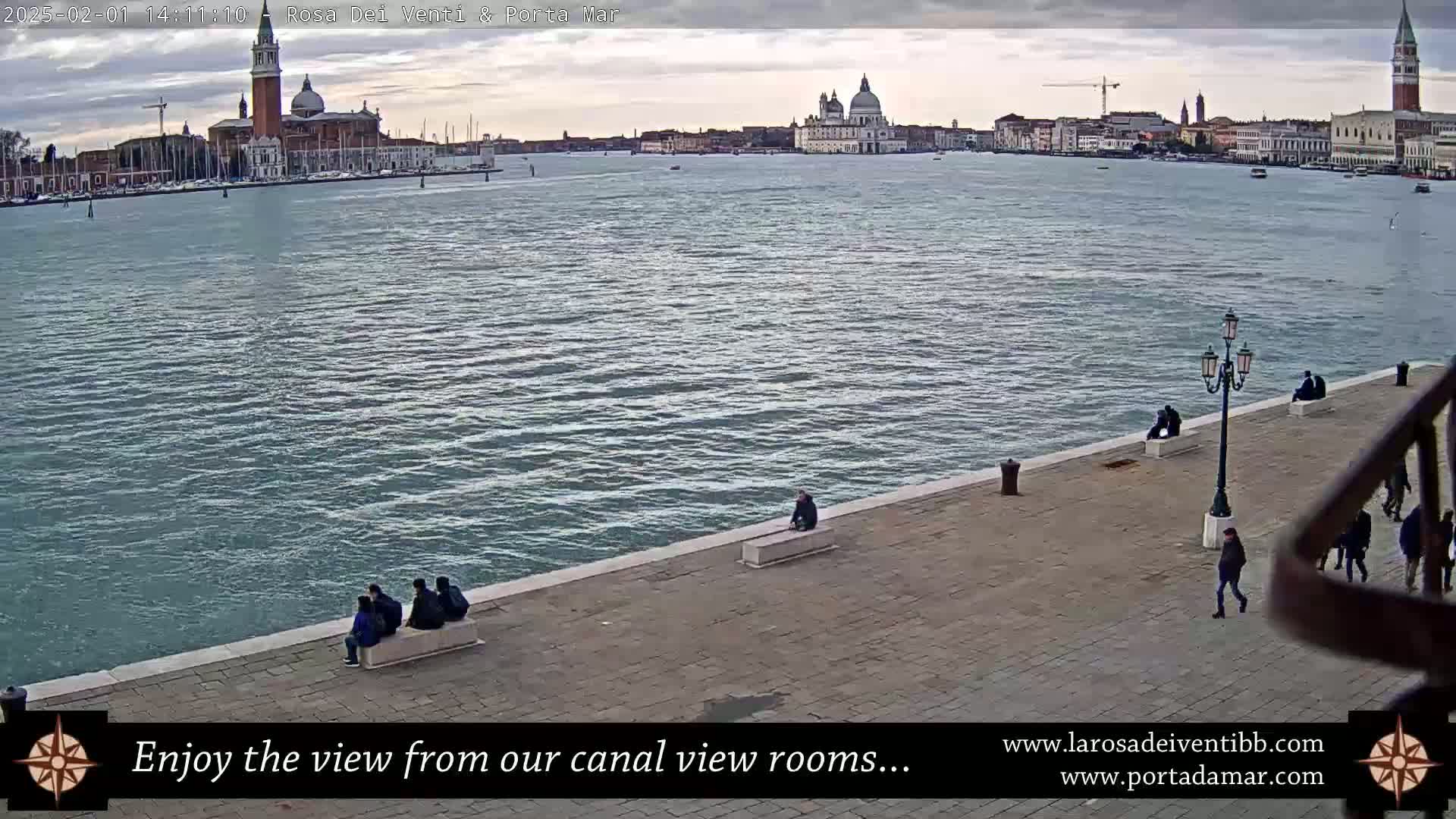 A wide shot shows a calm body of water with a city skyline in the distance under an overcast sky; several people are sitting and walking along a paved waterfront.