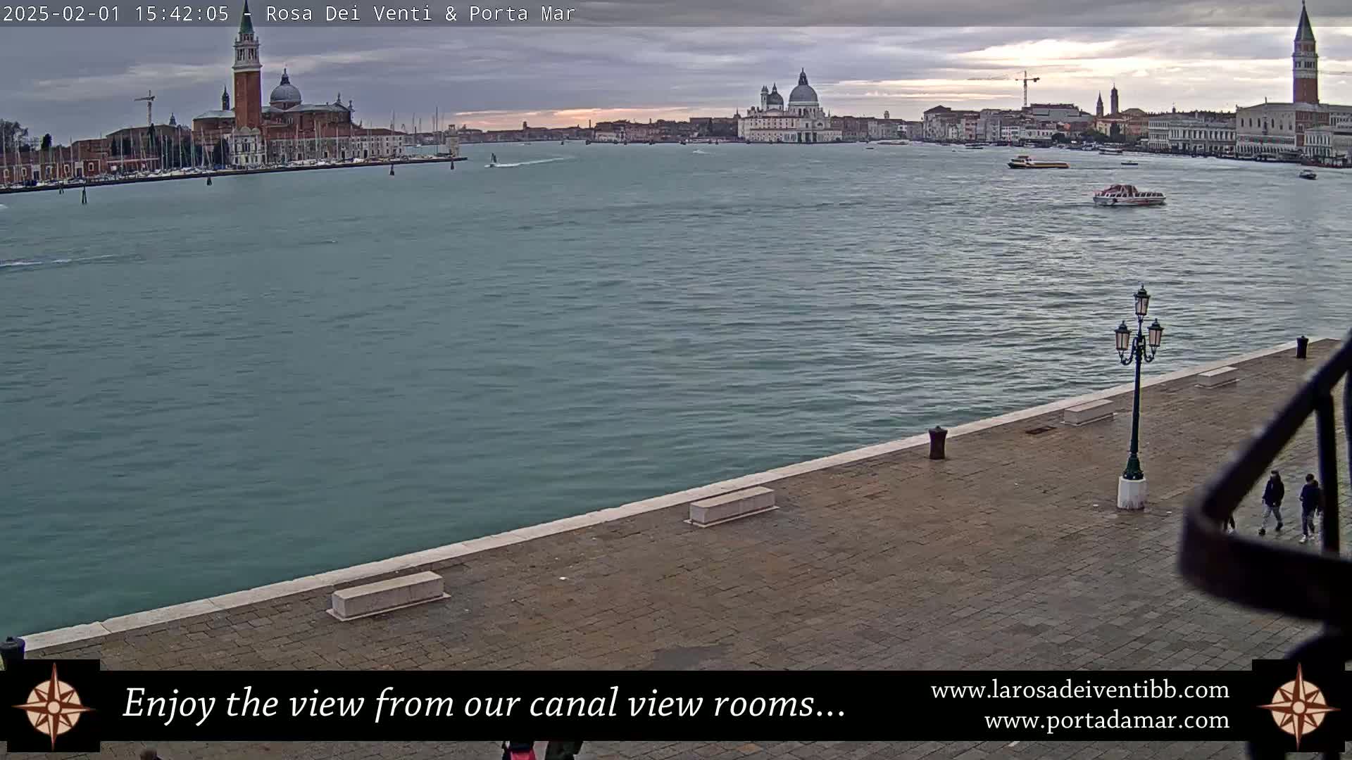A waterfront view of a calm canal in Venice, Italy, under an overcast sky, shows a cityscape with several prominent buildings, including a large church with a dome.