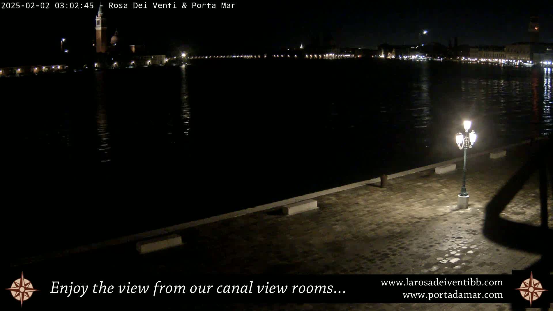 A nighttime view of a canal, with a city skyline faintly illuminated in the distance, and a single lamp post casting a shadow on a paved waterfront.