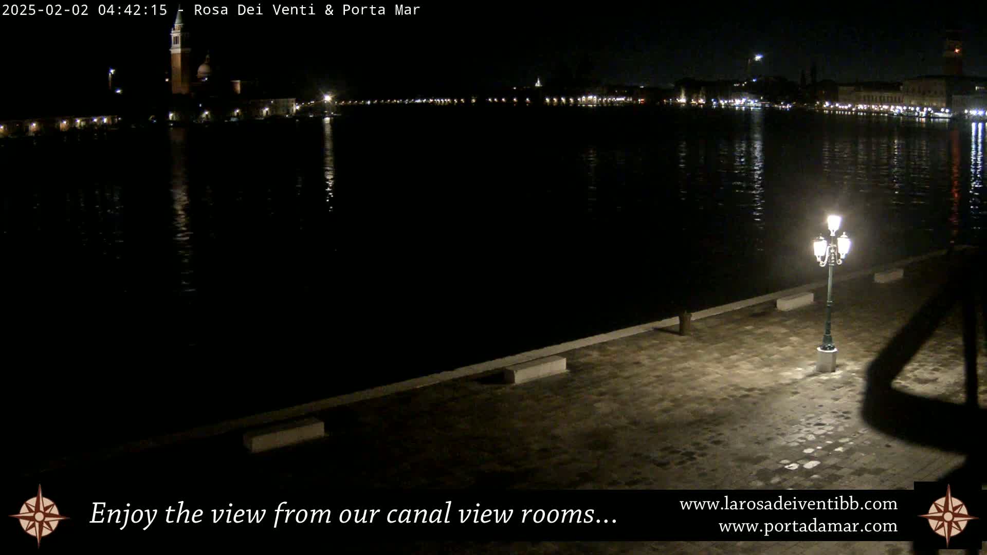 A nighttime view of a calm canal reflecting city lights, with a single lit lamppost on a paved walkway in the foreground.