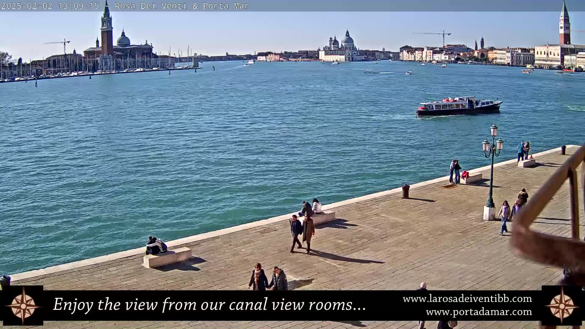 A sunny waterfront view shows a canal with boats and buildings in the distance, people walking along a paved walkway, and a few people sitting on benches.