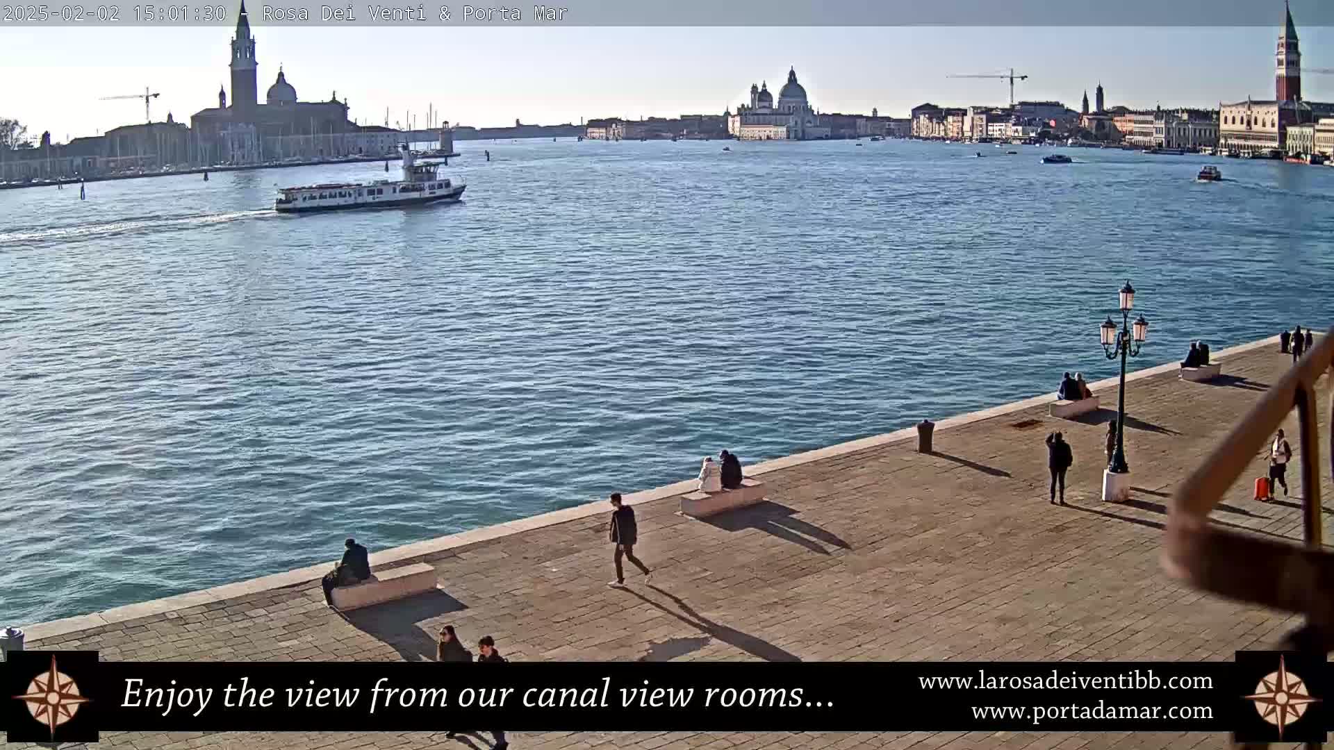 A sunny day in Venice shows a waterway with a boat and buildings in the distance, alongside a paved walkway with several people.