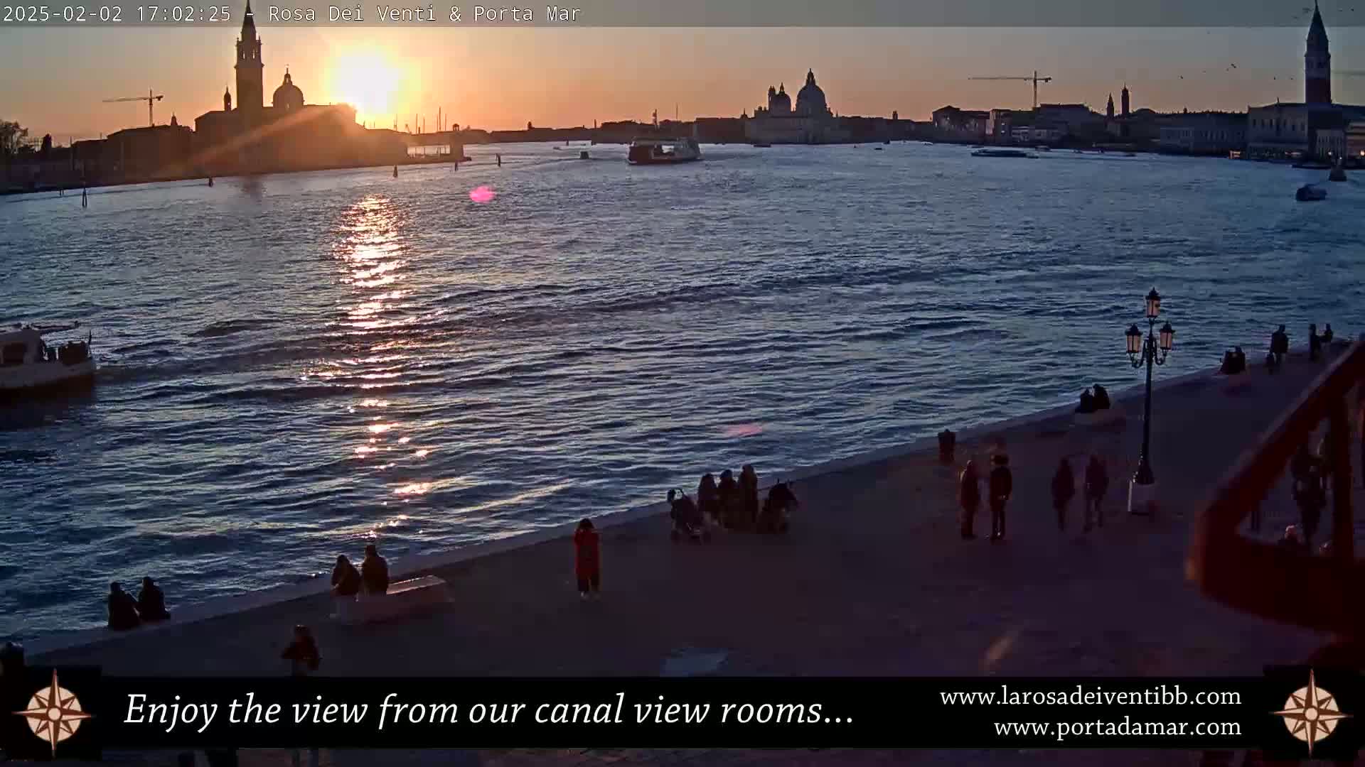 A sunset casts golden light on the water of a canal, as people stroll along a waterfront walkway overlooking a city skyline with prominent churches and a boat in the distance.