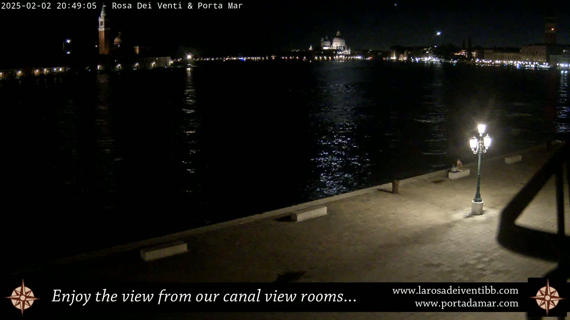A nighttime view of a canal with city lights across the water, a single lit lamppost on a paved waterfront, and a calm, dark-reflective surface.
