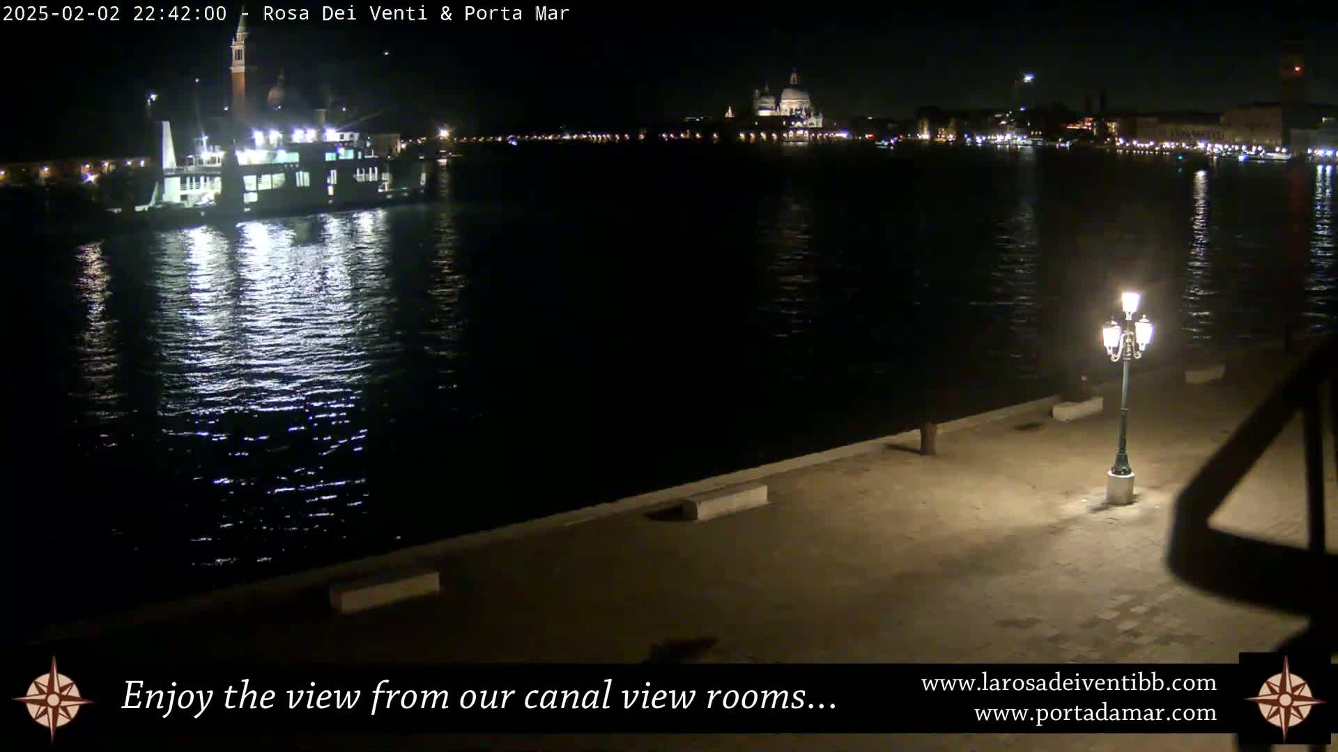 A nighttime view of a canal with a boat and city lights across the water, illuminated by a single lamppost on a paved waterfront.