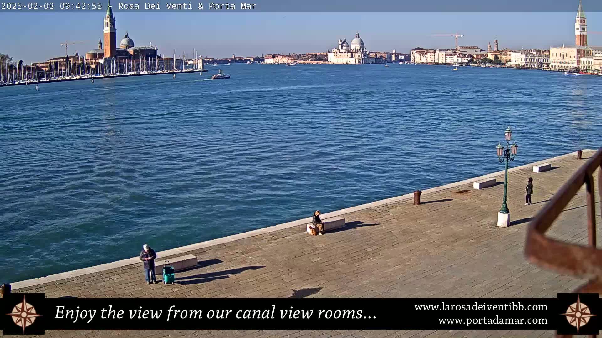 A sunny day shows a canal view of Venice, Italy, with several people walking along a stone embankment.