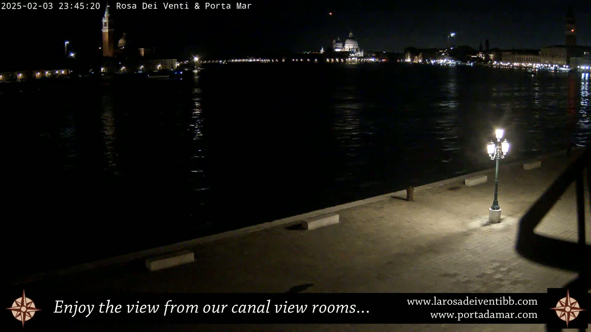 A nighttime view across a calm canal shows a cityscape with illuminated buildings and a single lit lamppost on a paved waterfront.