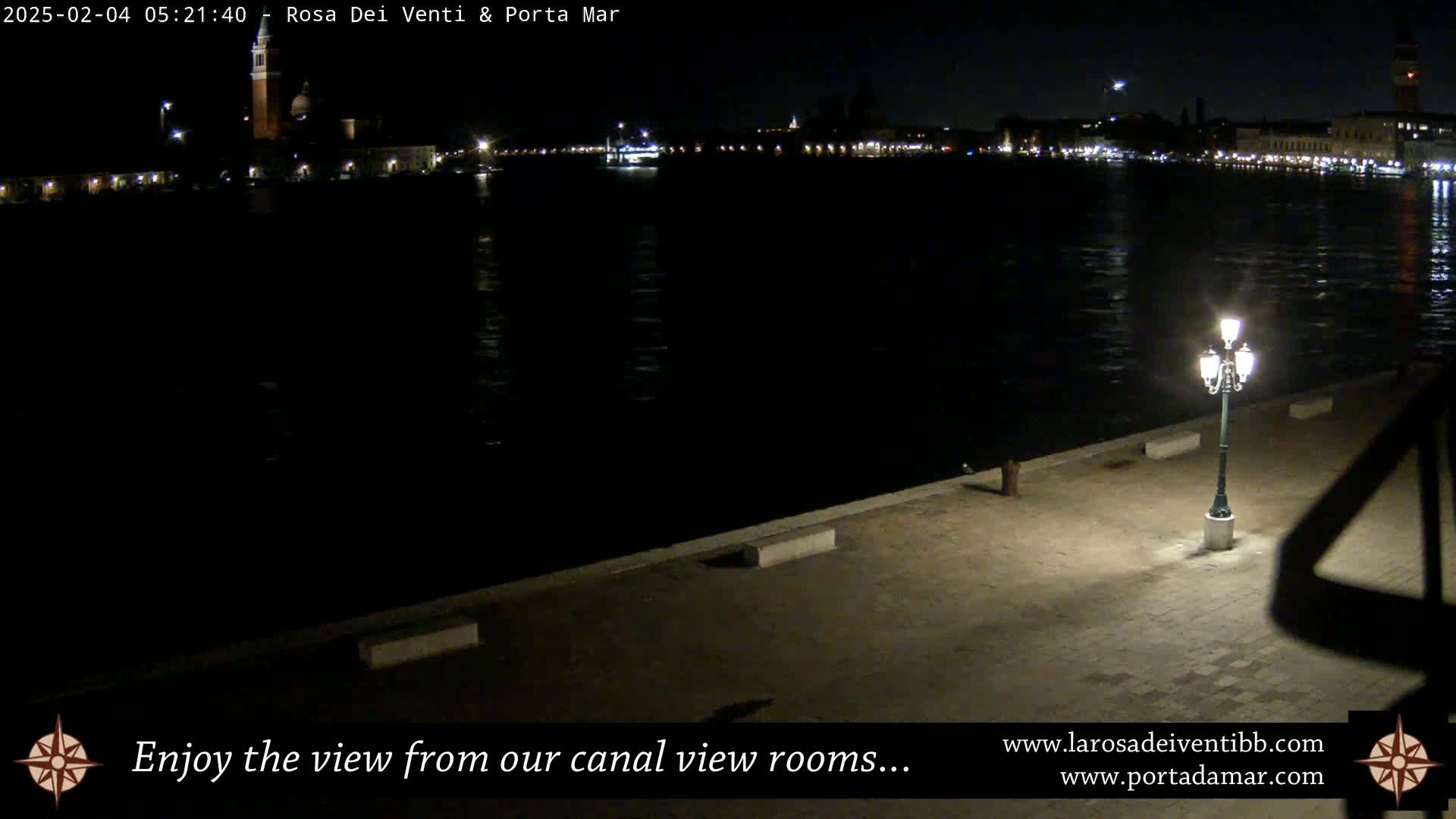 A nighttime view from a waterfront promenade shows a dark canal reflecting the lights of a distant cityscape.